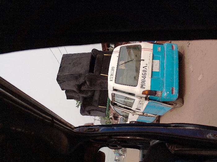 The image captures a street scene in Orie Uratta, Nigeria, viewed from within another vehicle, with its dark interior framing the left and right edges. A white and light blue commercial van, marked with "ICT/MCCRD" and "IM00460A" on its rear, is central. Two dark, upholstered armchairs with gold-colored leg accents are stacked on the van's roof. The van's rear passenger door is open, extending outwards. Its license plate holder displays "01U-162." The road is unpaved, consisting of reddish-brown dirt with scattered debris. In the blurry background to the left, other vehicles, potentially motorcycles, and indistinct figures of people are visible, suggesting activity on a thoroughfare. Buildings with corrugated roofs and some sparse foliage are present along the street. The sky is uniformly overcast.