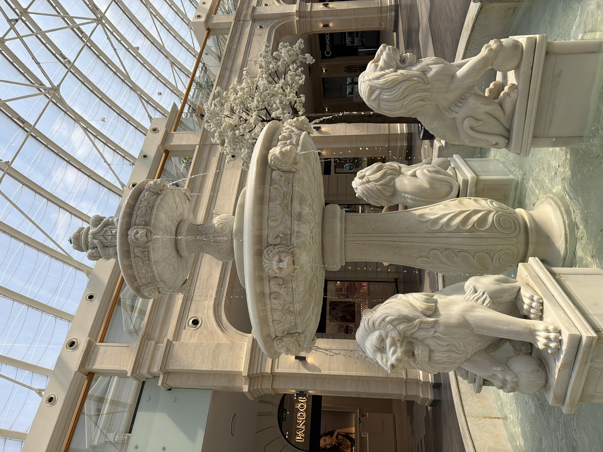 An elaborate marble fountain featuring three lion sculptures is centrally positioned within a brightly lit indoor atrium. The fountain's multi-tiered design includes ornate carvings of lion heads from which water cascades. The lions, depicted in a seated posture, each rest a paw on a sphere.
The atrium has a high, arched glass ceiling supported by a light-colored metal framework, allowing ample natural light to flood the space. The surrounding architecture is characterized by smooth, beige stone cladding and elegant columns. In the background, shops with illuminated signs are visible, including "PANDORA" and another with Cyrillic text that translates to "CEKУНДO". A decorative artificial tree with white blossoms stands behind the fountain. The floor is tiled in a light color. The image captures the fountain from a slightly low angle, emphasizing its grandeur.
The scene appears to be located in a public space such as a shopping mall or a large indoor complex. Considering the Cyrillic signage, a plausible location is a modern shopping center in Kyiv, Ukraine. No individuals are prominently featured in the image, with only a glimpse of a person in a display at the Pandora store and obscured figures in the background. The water in the fountain is clear and reflects the surrounding light.