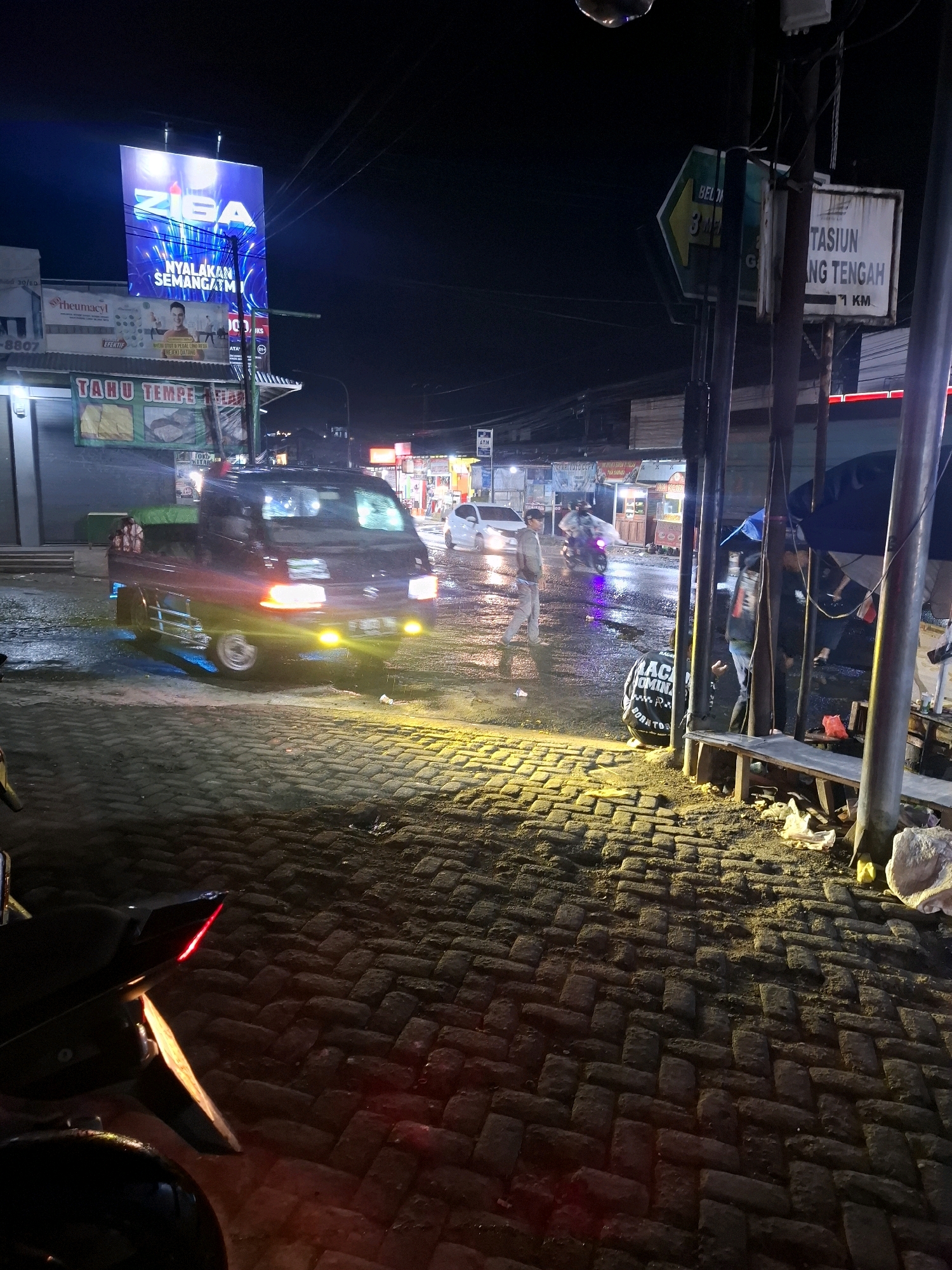 The image depicts a nighttime street scene in Ciheulang Tonggoh, Indonesia. The road is paved with dark bricks and is wet, suggesting recent rain. A dark-colored pickup truck is driving down the street, its headlights and auxiliary yellow lights illuminating the road ahead. To the right of the truck, a man in a light-colored shirt and pants stands on the wet road, appearing to be observing something. Further down the street, other vehicles and pedestrians are visible. A large digital billboard with bright blue and white lettering is prominently displayed on the left side of the image, advertising "ZIBA" and "NYALAKAN SEMANGATMU". Several other signs and banners are also visible, including one that reads "TAHU TEMPE MASAK" and another with a green arrow pointing to the left. The environment appears to be an urban or semi-urban area with various buildings and businesses lining the street. The overall atmosphere is one of a bustling night market or commercial district.
