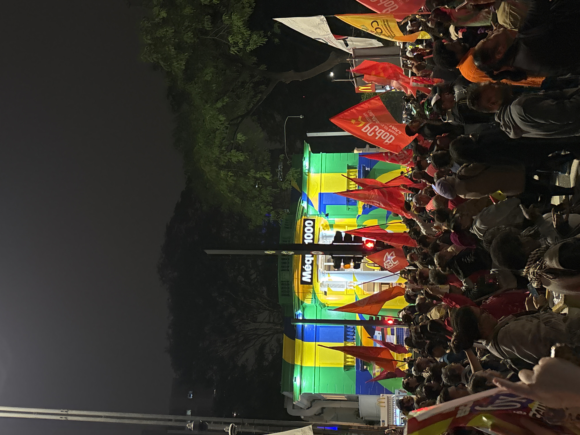 A nighttime scene in the city of São Paulo, Brazil, features a large crowd gathered in front of a building brightly painted in green, yellow, and blue horizontal stripes. The building has signage in Portuguese "MéQ1 1000." The crowd holds numerous red and other colored flags, and some people are looking up or gesturing. Trees are visible on the left side of the frame, contrasting with the urban environment. Streetlights illuminate the scene, with some people visible in the foreground. The presence of flags suggests a demonstration or gathering of political or social significance.