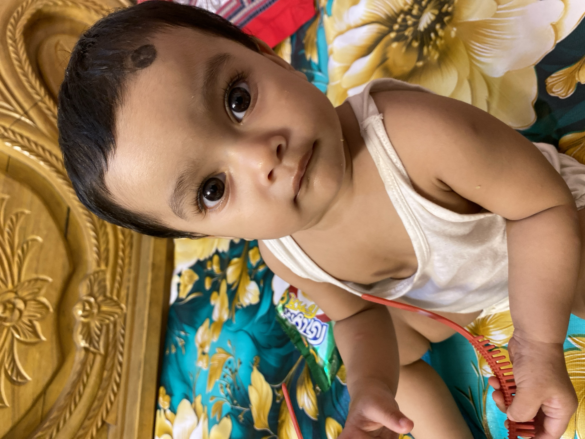 A baby with dark hair and brown eyes is looking up at the camera. The baby is wearing a white tank top and holding a red comb. The baby has a dark spot on its forehead. The baby is sitting on a bed with a floral pattern. There is a wooden headboard on the left side of the image. A packet of snacks is visible in the background. This image was taken in Dhaka, Bangladesh.