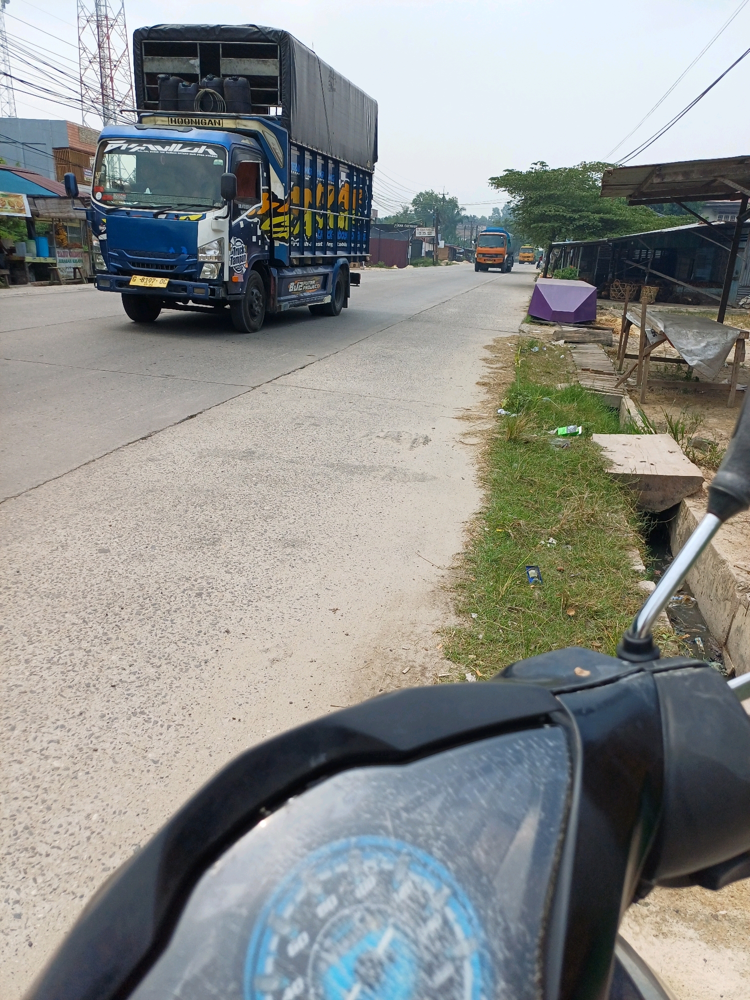The image depicts a roadside scene in Dumai City, Indonesia, likely on a slightly overcast day. In the foreground, the handlebars and instrument cluster of a black motorcycle are visible, suggesting the photo was taken from the rider's perspective. The road itself is paved, with a textured section to the right and a smoother section to the left.
Dominating the mid-ground is a large, blue and white truck with a black tarp-covered cargo bed. The truck has various decals and markings, including the word "HOONIGAN" on the top of the cab and "PUTRA PROJECT" on the side. It is traveling on the road. Further down the road, two smaller orange trucks are visible in the distance, also in motion.
The environment along the roadside includes a drainage ditch with overgrown grass and scattered debris. On the right side of the road, there are several makeshift structures, including wooden stalls with covered tops and what appears to be a purple tarp-covered object. Buildings and utility poles are visible in the background, hinting at a semi-urban or commercial area. The overall impression is a typical street scene in a developing urban environment.