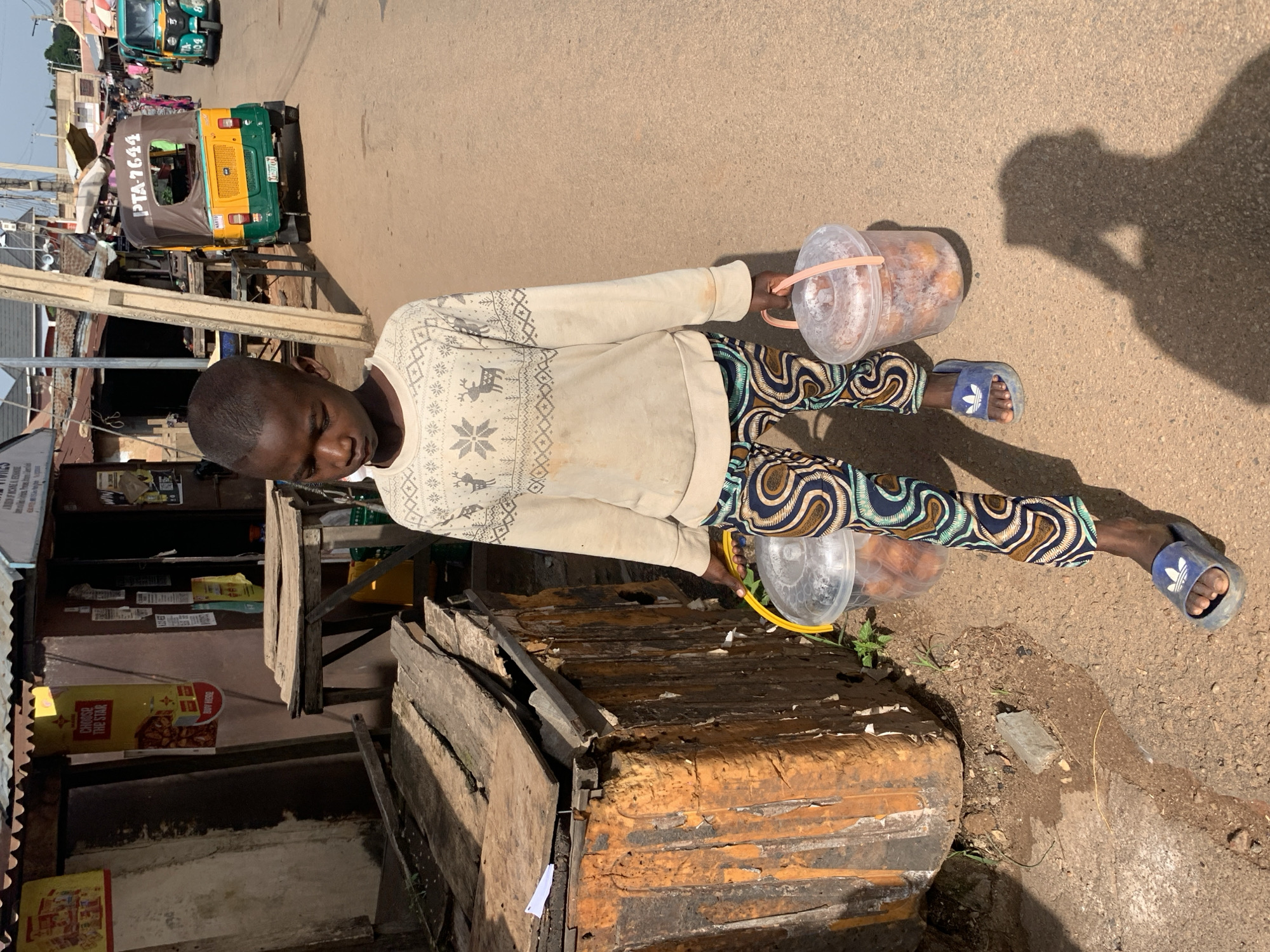 A young boy with dark skin, a shaved head, and a light-colored, long-sleeved shirt with a patterned print walks on a dusty, paved street in Jos, Nigeria. He wears colorful patterned pants and blue Adidas slides. In each hand, he carries a clear plastic bucket with a light pink handle. The buckets appear to contain fried food items. To the left of the boy is a dilapidated, orange-colored structure made of wood and corrugated metal, with stacks of planks next to it. In the background, a yellow and green auto-rickshaw with the license plate PTA-7644 is visible, along with other vehicles and market stalls. The scene suggests a busy street or market environment.