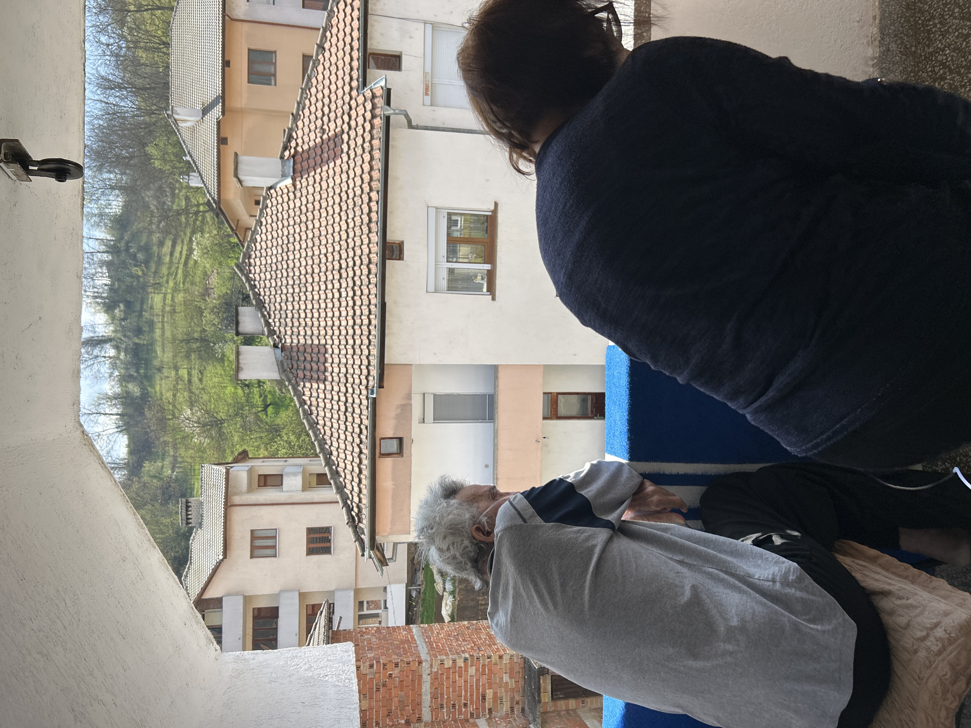 A slightly tilted medium shot shows two people looking out from a balcony over a densely built area. An elderly man with gray hair is sitting in the foreground, wearing a gray t-shirt with dark blue stripes on the shoulders and sleeves, and dark trousers. He is seated on a beige blanket and a blue-and-white striped blanket, and is positioned closer to the camera than the other person. The second person, standing behind the man, is wearing a dark blue sweater and has dark hair. Both figures are oriented towards the view from the balcony.
The background features a densely packed urban landscape. There are several buildings, mostly two or three stories high, with a variety of roof types, including tiled roofs. The building exteriors are a mix of materials, including painted walls, brick, and plaster. Some of the buildings are closely spaced. Further back, there's a green, treed hill, likely part of the surrounding landscape. The shot is taken from a slightly elevated viewpoint, suggesting a balcony or higher floor. The window is visible on the top left of the image. The location is likely Zlatograd, Bulgaria.