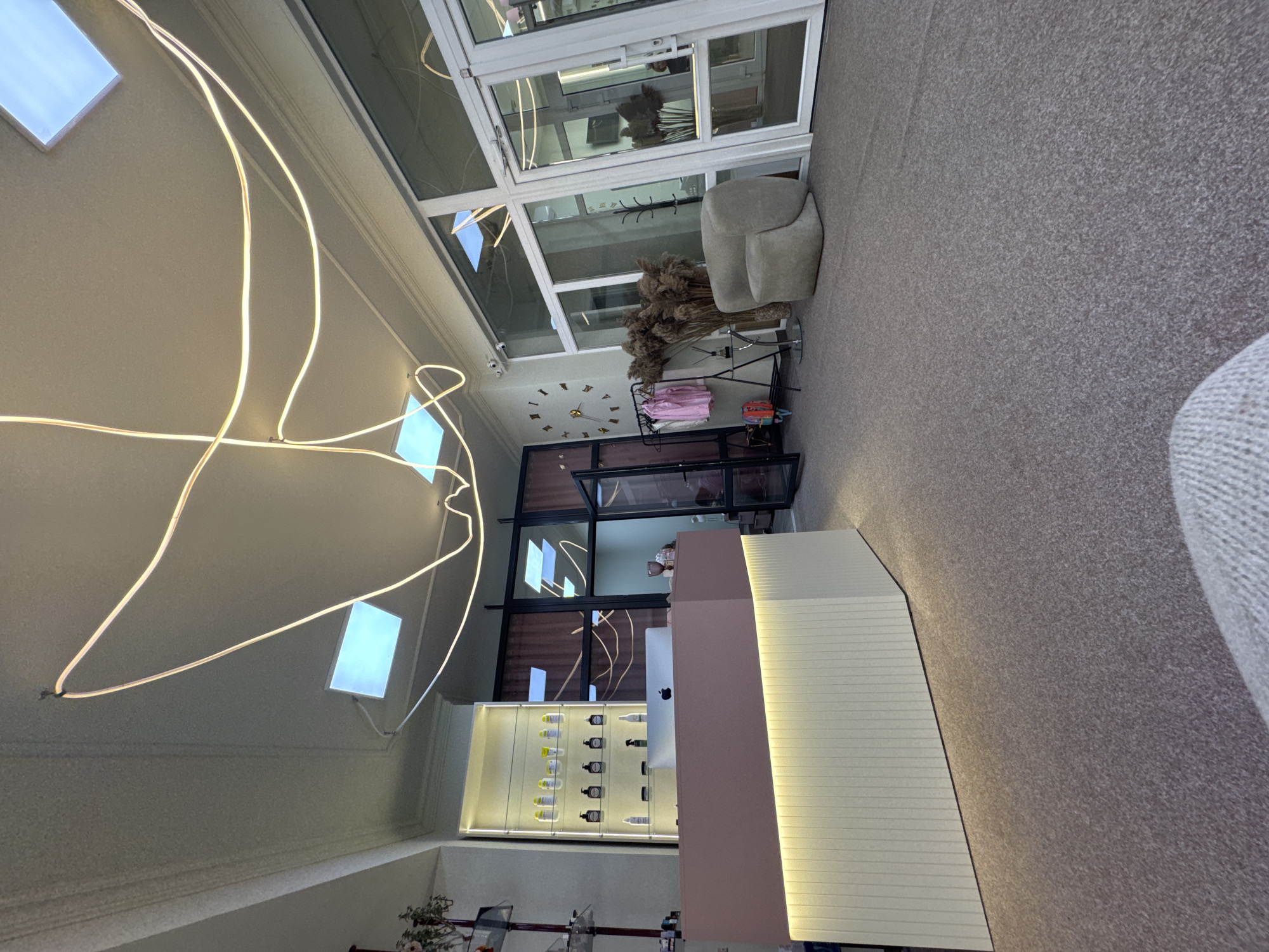 The image depicts the interior of a commercial establishment, likely a salon or boutique, located in Chernivtsi, Ukraine. The scene is characterized by a modern and minimalist aesthetic.
Dominating the foreground is a reception desk, a substantial, angular structure with a pale pink front and a ribbed, cream-colored base illuminated by soft lighting. A silver Apple monitor sits atop the desk. Behind the reception desk, a glass-fronted display cabinet showcases various bottles and containers, suggesting retail products. The walls are a light color, and the ceiling features recessed lighting panels and an elaborate, looping neon light fixture that appears to be a central decorative element.
To the right of the reception desk, a large wall clock with Roman numerals is prominently displayed. Nearby, a rack holds several items of clothing, primarily pink garments, indicating a retail aspect to the business. A unique, armchair-like seating unit with a rounded, organic shape is positioned to the right, accompanied by a tall arrangement of dried grasses.
The environment appears to be a well-lit interior space with large glass doors and windows, suggesting a connection to the outside or a spacious feel. The floor is covered in a textured, grey carpet. The overall impression is one of a clean, contemporary, and welcoming commercial space in Chernivtsi. There are no people clearly visible in the image.