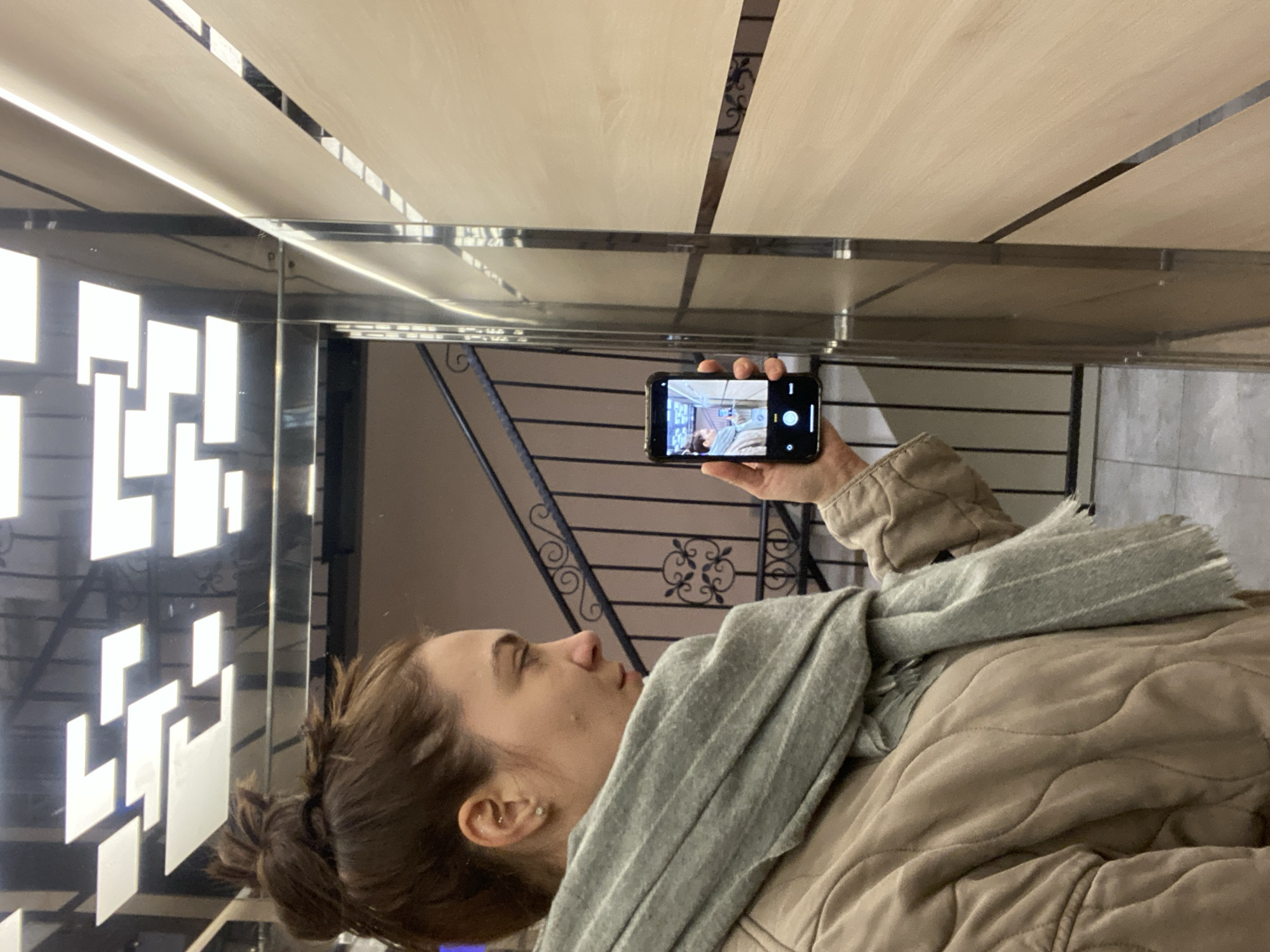 A woman is depicted in profile, facing towards the right side of the frame, in what appears to be a modern indoor setting. She has dark hair styled in a bun and wears a quilted beige jacket along with a light grey striped scarf wrapped around her neck.
She holds a black smartphone horizontally in her right hand, positioning it at eye level. The phone's screen is active, displaying a camera interface. Within the phone's display, a scene is visible, possibly a reflection of the surroundings or a previously captured image, which includes a person and light-colored wall panels. Typical camera controls, such as a white circular shutter button and a numerical "1" (likely indicating a setting or count), are visible on the screen.
The background features reflective surfaces, possibly glass or polished panels. To the left, a dark surface showcases a pattern of glowing white rectangular shapes, suggesting modern illuminated wall art. Behind the woman, an ornate dark metal railing with intricate scrollwork is visible, hinting at a staircase or balcony. Above, the ceiling consists of light-colored rectangular panels separated by darker lines, which are reflected in the glossy surfaces below. The overall environment is well-lit and has a contemporary design aesthetic, consistent with a public building or commercial space in Novi Sad, Serbia. The woman appears focused on her phone.