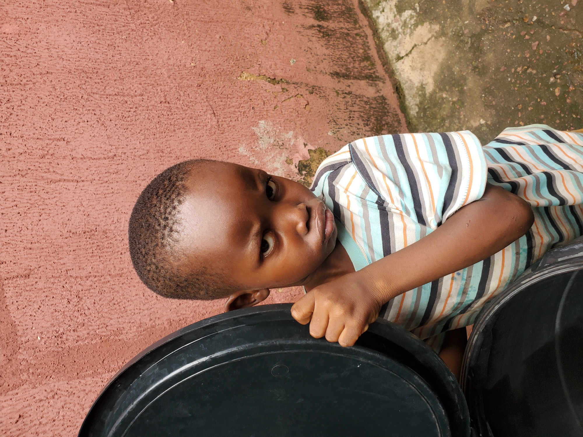 The image features a young child, likely a boy, leaning over two large black plastic containers. He is positioned in a way that suggests he is resting or playing, with his right hand gripping the edge of the closer container. The child is wearing a striped t-shirt with horizontal bands of blue, white, orange, and black. His expression is somewhat serious, with his eyes looking directly at the camera.
The background consists of a textured, reddish-brown wall, possibly a building exterior, with some patches of lighter, possibly mossy or weathered concrete. The ground appears to be a mix of concrete and possibly dirt or gravel, with some discoloration and evidence of dampness. The lighting suggests it is daytime, but the specific time is not discernible.
The setting appears to be outdoors, consistent with the visible ground and wall textures. Given the location context of Jos, Nigeria, the scene likely depicts a common residential or communal area. There is no visible text in the image. The overall impression is a candid, everyday moment captured with a slightly tilted perspective.