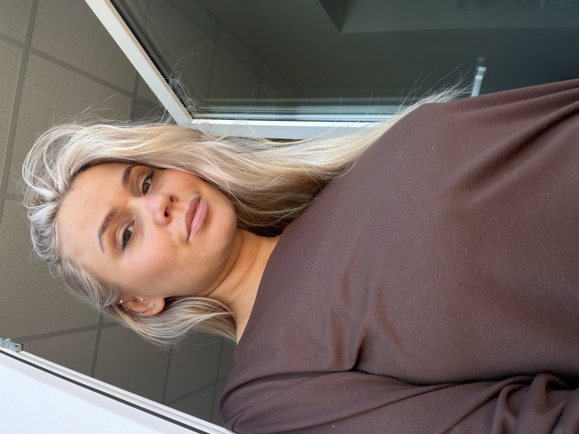 This close-up image, taken in Sarapul, Russia, features a young woman looking directly at the camera with a neutral, slightly pouting expression. Her head is tilted, and the shot appears to be a high-angle selfie. She has long, blonde, slightly wavy hair that frames her face, fair skin with subtle freckles, and light-colored eyes. Her left ear is adorned with multiple piercings, including a silver hoop, several small studs, and a delicate chain. She is wearing light makeup, accentuating her lips with gloss.
The woman is dressed in a plain, long-sleeved top or dress in a dark brown or mauve color, made from a soft, presumably comfortable fabric.
The background suggests an indoor setting during the daytime, with natural light illuminating the scene. A white window frame is visible above her, and through it, a grey ceiling with a grid pattern, characteristic of an office or commercial building, can be seen. Another white structural element is visible near her left shoulder. No text is visible in the image.