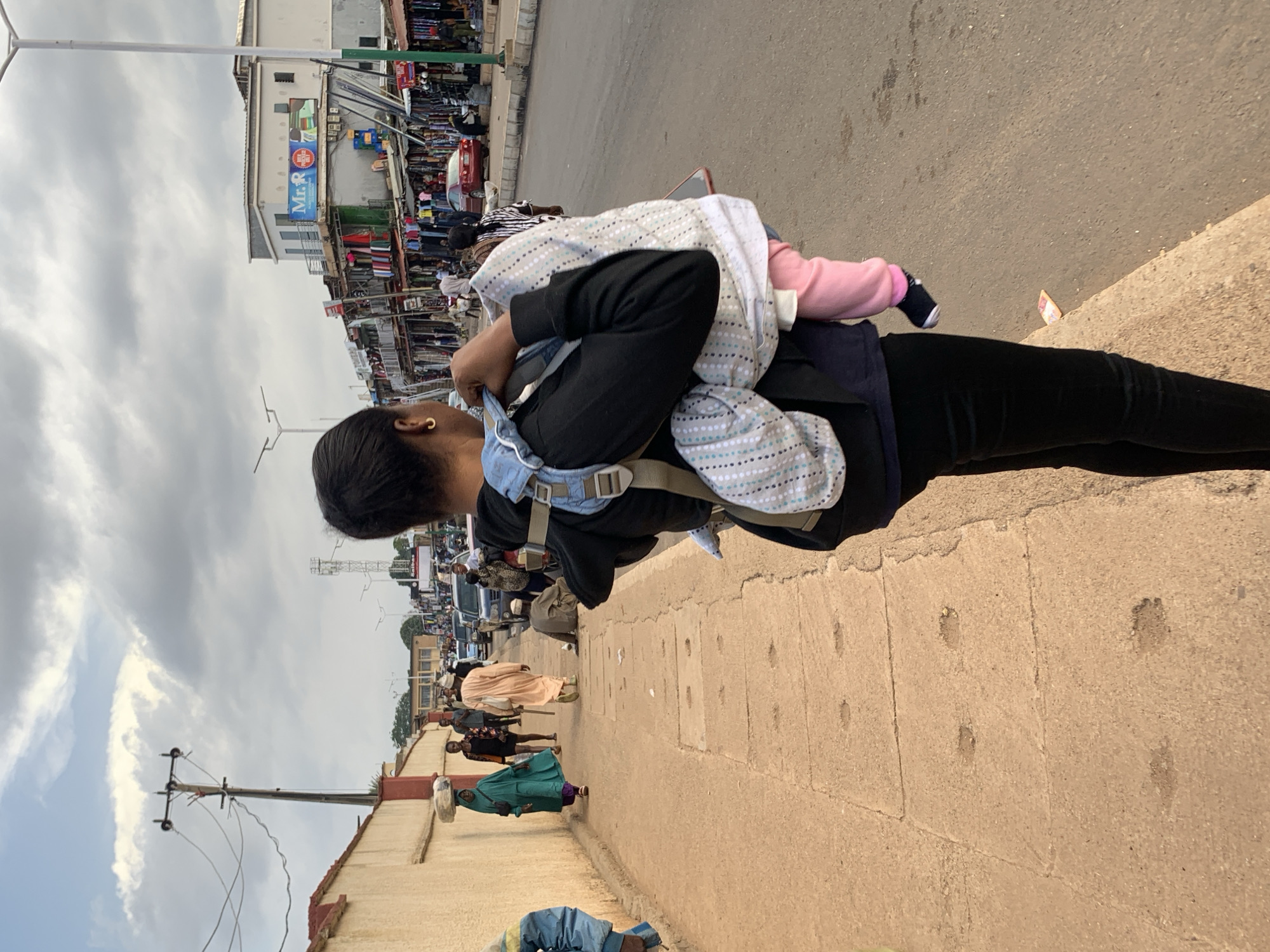 In a street market scene in Jos, Nigeria, a woman is seen from behind carrying a baby in a fabric carrier. The woman is wearing a black top and dark pants. The baby is wrapped in a blanket and appears to be sleeping or resting. The background shows a busy street with various shops and stalls lining the road. People are walking and going about their day. A red car is parked on the side of the road. Power lines and poles are visible overhead. The sky is overcast, suggesting a cloudy day. The scene depicts a typical day in a bustling market environment. There is a sign with "Mr. R" visible on one of the buildings, which may be a business name. The overall impression is one of everyday life in a vibrant urban setting.