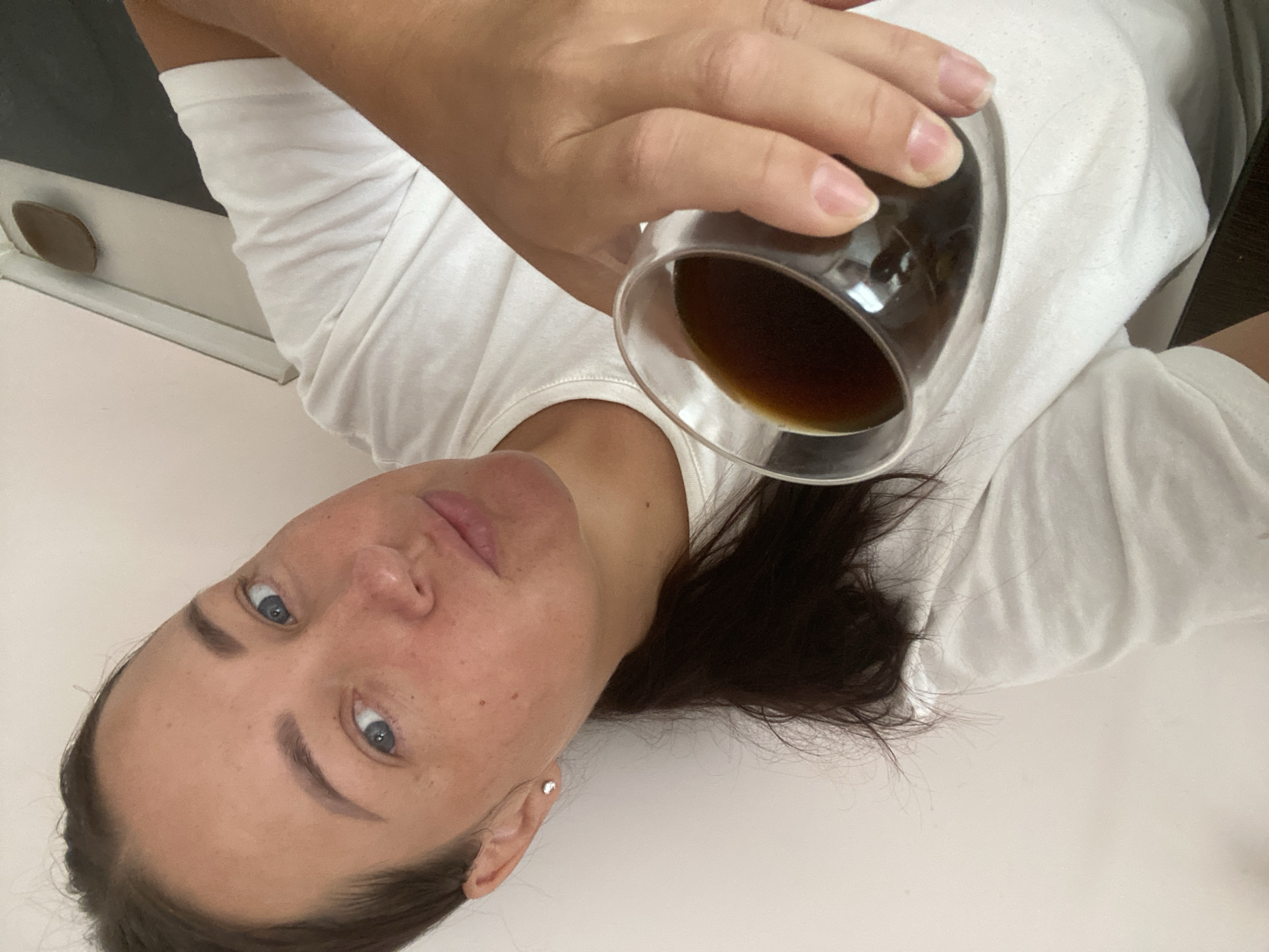 A woman with dark brown hair pulled back in a ponytail, wearing a white t-shirt, holds a clear glass containing a dark liquid, likely coffee, towards the camera. She has blue eyes and a fair complexion with freckles. Her left ear has a small stud earring. The background consists of a light-colored wall and a dark surface, possibly a countertop or table. The image is a close-up selfie.