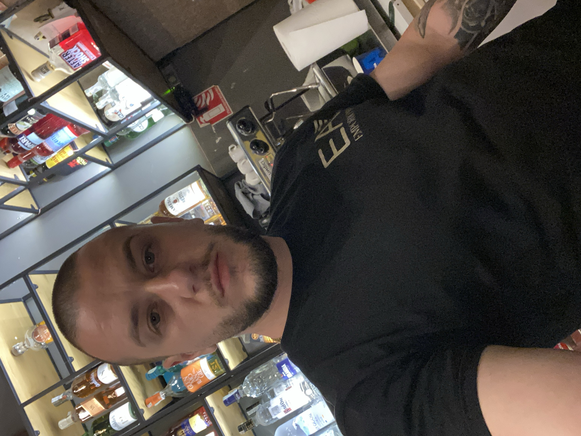 The image shows a man taking a selfie in front of a bar in an unidentified location. A man with a shaved head and short, dark beard is centered. He is wearing a black t-shirt with gold letters on the upper right chest. The bar behind him displays various alcohol bottles arranged on shelves. A coffee machine is visible on the bar top. The background includes bar equipment and supplies. The location or city is not discernible from the image.