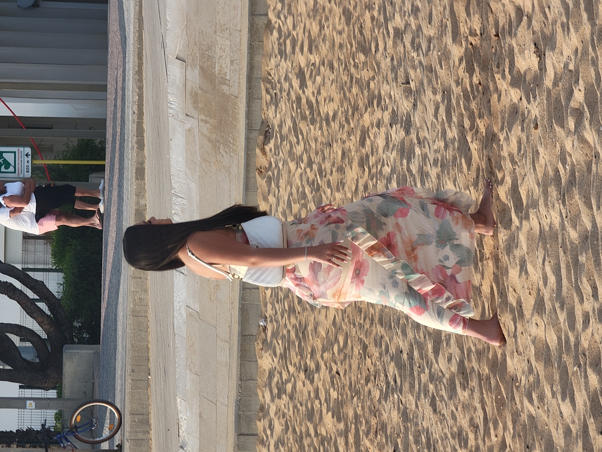 A woman with long dark hair is walking barefoot on a sandy beach in Quarteira, Portugal. She is wearing a flowing, floral-patterned maxi dress and carrying a white purse over her shoulder. Her right arm is bent at the elbow and her hand rests on her hip, revealing a bracelet and a ring on her finger. In the background, a bicycle is parked near a tree and a paved walkway. Further in the distance, two people stand near a sign that reads "CAMINHO DE EVACUAÇÃO EVACUATION ROUTE 820 m". The beach sand is textured with ripples and shadows, indicating the time of day is likely late afternoon or early evening.