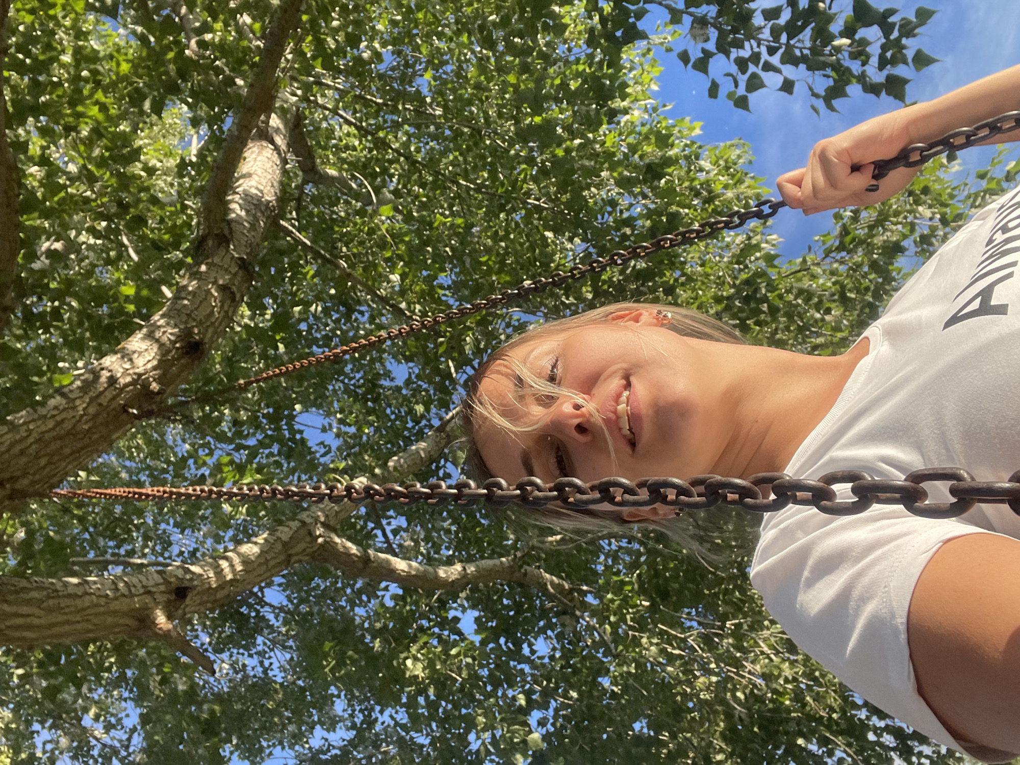 A low-angle, inverted shot captures a smiling young woman on a swing, looking directly at the camera. Her blonde hair is partially visible and slightly wind-blown around her face. She is wearing a white t-shirt, on which a dark, partial letter "A" and other indistinct text are visible. Her right hand grasps one of the two thick, rusty metal chains that are prominently featured, stretching horizontally across the frame.
The setting is outdoors, beneath a canopy of lush green trees, likely in a park or recreational area in Chaikovsky, Russia. Bright sunlight filters through the leaves, illuminating the textured bark of the tree trunks and branches. A clear blue sky is visible between the foliage and above the woman. The vibrant lighting suggests it is daytime, probably midday or afternoon, under sunny weather conditions. The woman's broad smile conveys a sense of joy and playfulness, indicative of enjoying the activity of swinging.