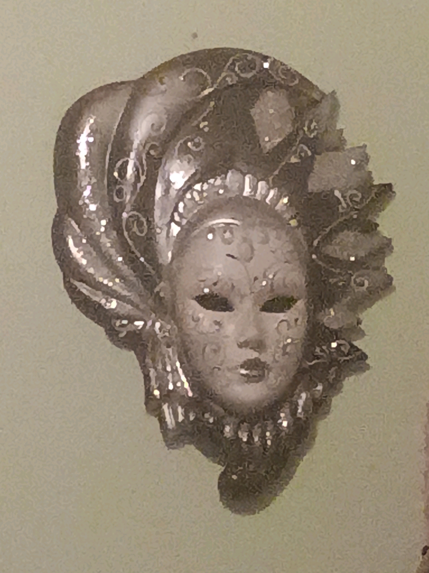 This is a close-up, slightly out-of-focus, head-and-shoulders shot of a decorative mask. The mask is silver-toned and features intricate detailing. It has a headdress with feathery or petal-like protrusions and ornate swirling patterns on the mask itself. The background is a plain, light green textured surface, possibly a wall. There is no indication of the city of Varallo Pombia, Italy in the image.