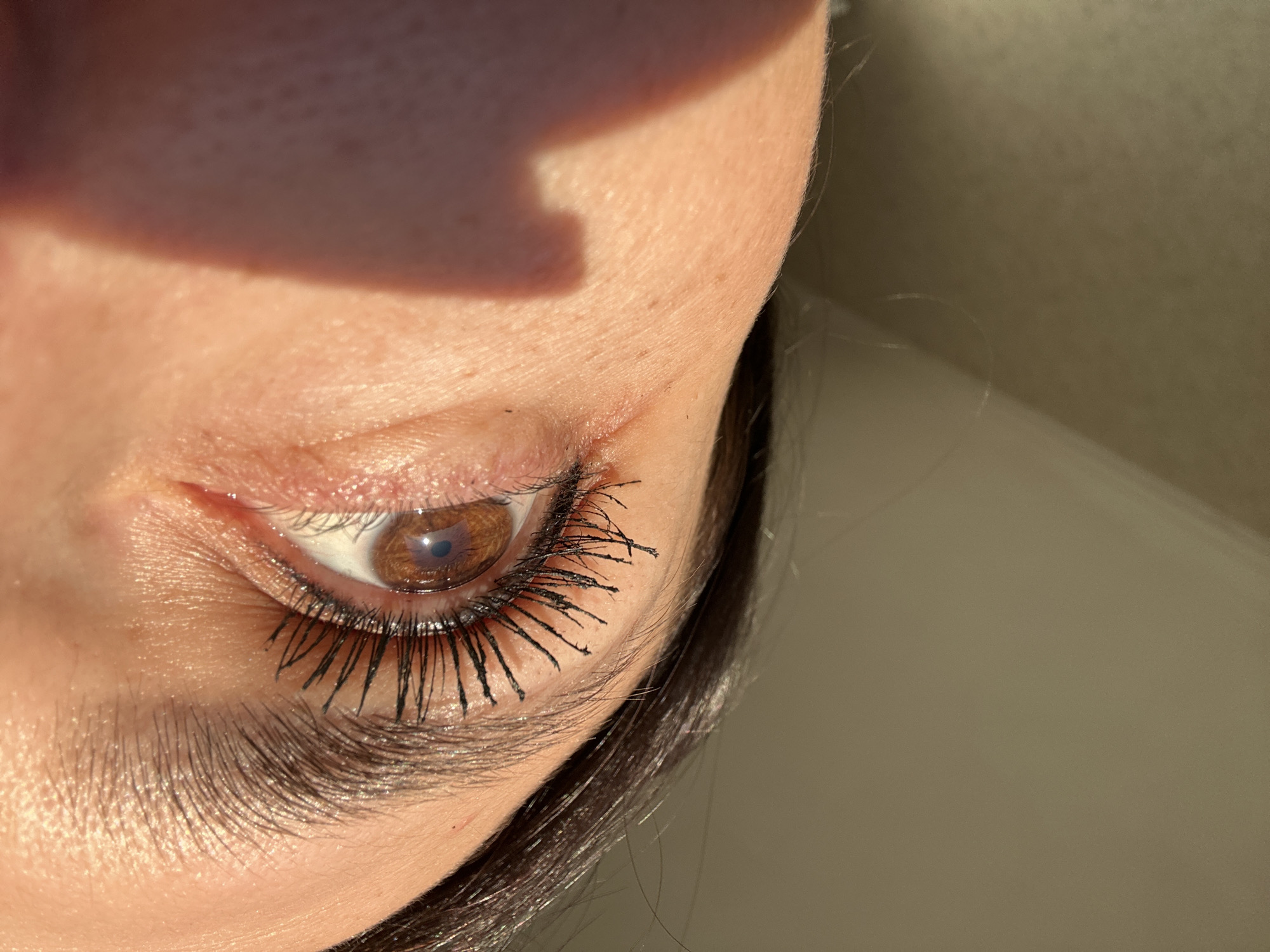 A close-up, high-angle shot shows a person's left eye and surrounding facial features. The eye is open, with brown iris and a discernible pupil. The person has applied mascara, resulting in defined and somewhat clumpy eyelashes. The surrounding skin is light-toned, and some wrinkles are visible around the eye. A dark shadow is cast on the upper part of the face near the eyebrow. Dark hair is seen near the ear. The background is a neutral, possibly light-colored wall or surface. The exact location is not discernable, and there is no indication it is in a city.