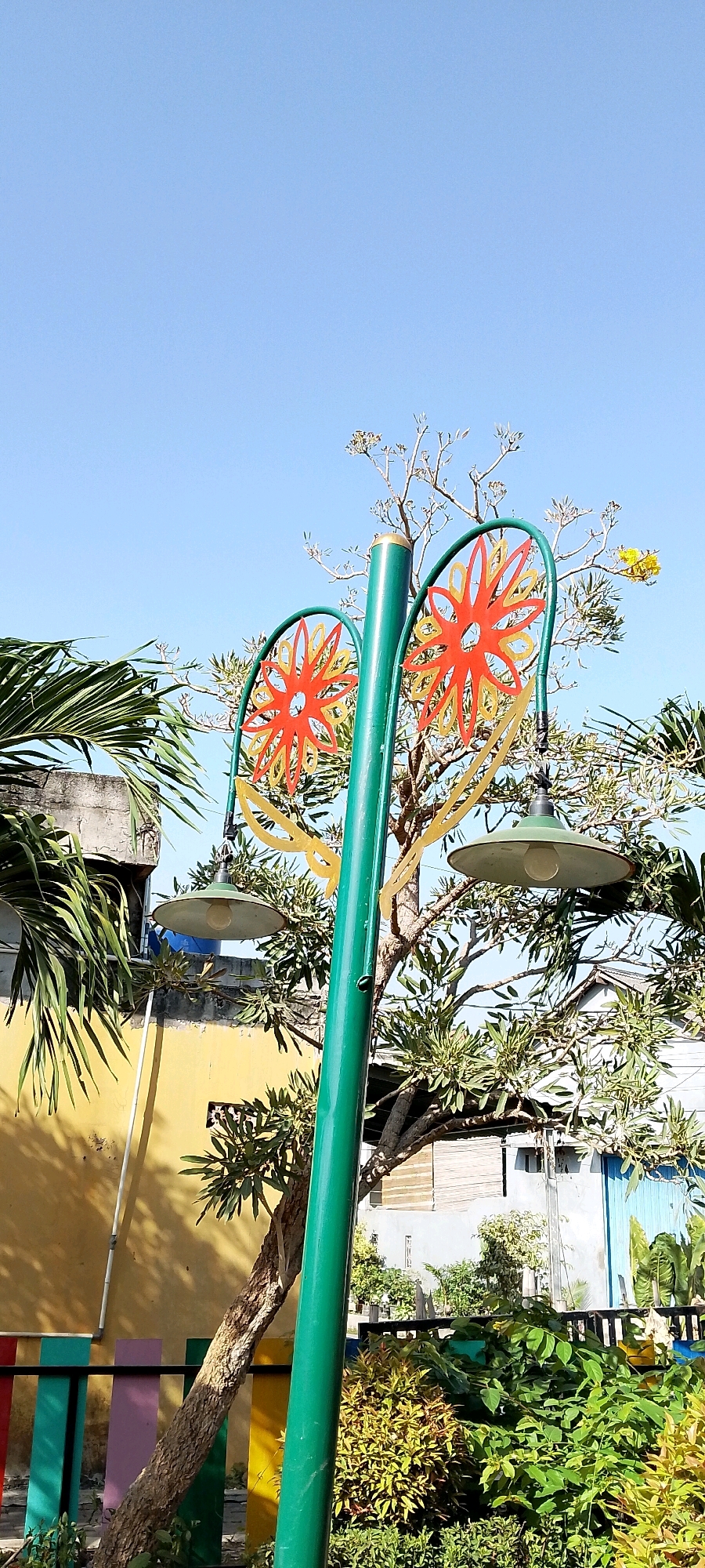 A green lamppost with decorative orange and yellow flower-like accents stands in a garden under a clear blue sky. The lamppost has two green lampshades, each with a visible light bulb. In the background, a yellow building is partially visible, along with trees, shrubs, and a colorful fence. To the left of the lamppost, a palm frond extends into the frame. The scene appears to be an outdoor public space, possibly a park or a street in Satriamekar, Indonesia. There are no people or animals present in the image.