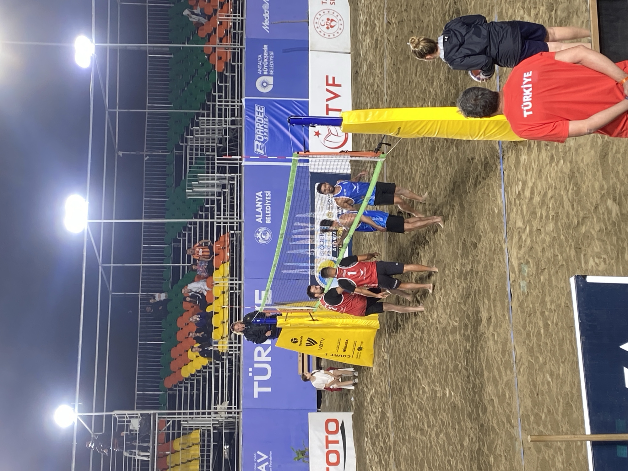 The image depicts a beach volleyball match, likely held in Alanya, Turkey, based on the banners and signage in the background. Two teams, one in red and black, and the other in blue and white, are positioned on either side of a net. A person wearing a red shirt with "TÜRKİYE" printed on the back is standing near the net on the right side of the image. Another person in a black jacket is standing near the person in the red shirt. The court is surrounded by spectator seating, with some people in the stands. Various sponsorship and branding elements are visible, including logos of "VBTV", "MIKASA", "TVF", "ALANYA BELEDİYESİ", and "BOARDEE SPORTS", along with the word "TÜRKİYE" prominently displayed on a blue banner. The event appears to be taking place at night, illuminated by stadium lights. The ground is composed of sand, and there is a scorer's table with a person sitting at it near the barrier.