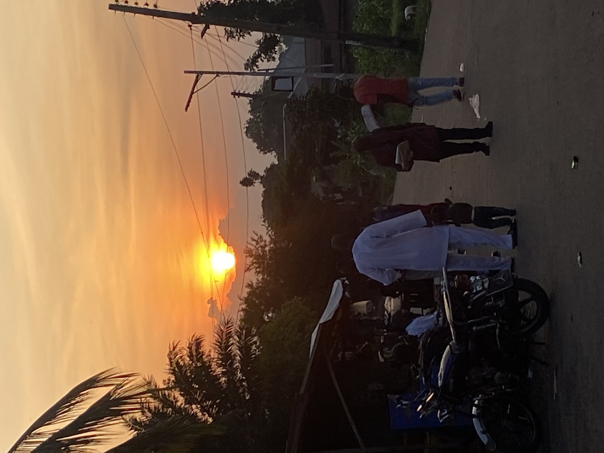 This image captures a sunset scene in Nguzu Edda, Nigeria, depicting a bustling street environment during the golden hour. The sky is dominated by intense orange and yellow hues, with the sun prominently visible on the horizon, partially obscured by dark cloud formations. Dense tropical vegetation, including large palm fronds on the left and various trees and bushes, is silhouetted against the bright sky, primarily on the left and in the background.
On the left side of the frame, a makeshift market stall or small shop with a dark roof and a white canopy is visible, where several indistinct figures are gathered. Parked on the road near this stall is a dark-colored motorcycle, likely blue or black, featuring chrome fenders and a visible license plate frame, oriented towards the right.
The central and right portions of the image feature a dark, likely asphalt or packed dirt road. Along the right side of the road, multiple wooden utility poles with an array of power lines and a street light fixture extend vertically.
Several individuals are present on the road, walking towards the right. In the midground, a person wearing a white, traditional long-sleeved garment and trousers is walking, accompanied by a small child in dark clothing walking beside or slightly behind them. To their right, another individual in dark clothing is walking while holding a rectangular object resembling a tray or container. Further right, a person in a red shirt and blue jeans is also walking away from the viewer. Scattered debris, including a crumpled white object and a small dark item, is visible on the road surface. In the far right midground, a small, light-colored dog is observable near the edge of the road. Distant structures and more vegetation are faintly visible further down the street on the right.