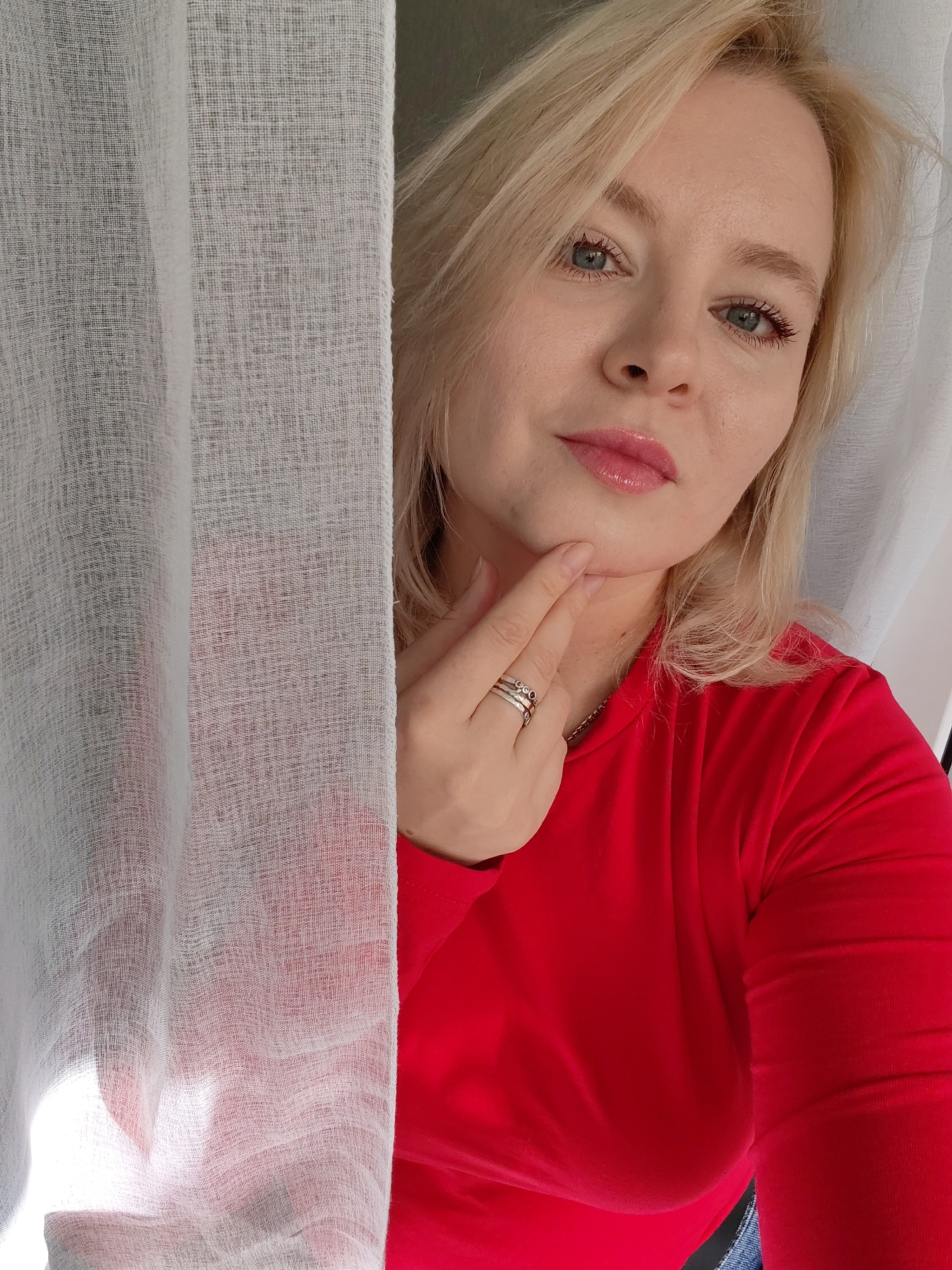 A woman with blonde hair and blue eyes, wearing a red top and rings, is positioned behind a sheer white curtain. She is touching her chin with her hand. The image appears to be a selfie taken indoors. There is no visible information in the image to indicate its location in Izhevsk, Russia.