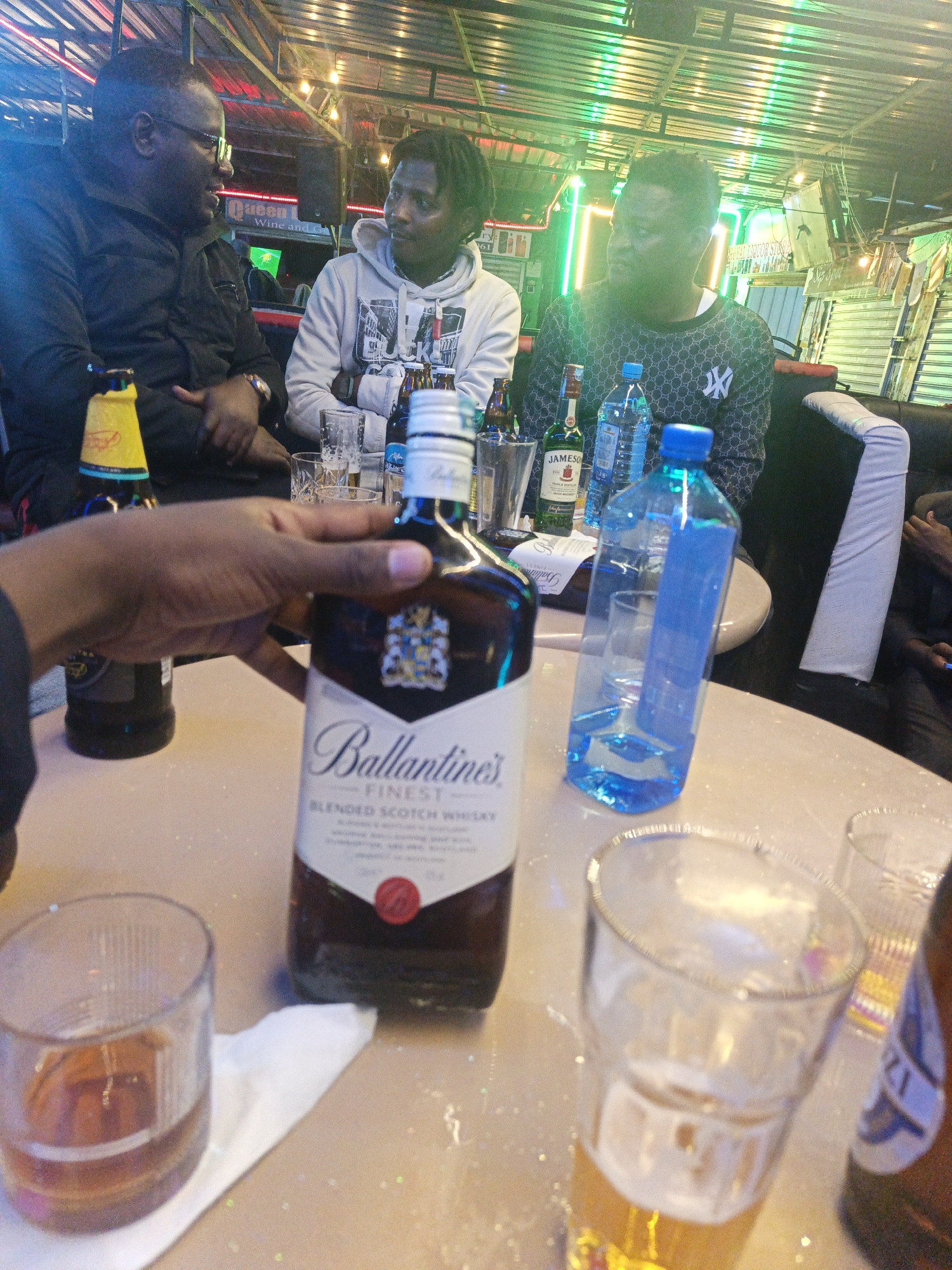 The image captures a casual social gathering of three men at a bar or lounge in Nairobi, Kenya, during the night or late evening.
In the foreground, a hand holds a prominent bottle of Ballantine's Finest Blended Scotch Whisky. To its left, a glass contains an amber liquid, likely whisky, resting on a white napkin. Further left, a yellow-capped beer bottle is visible. On the right, a tall glass filled with beer sits on the table, along with another partially visible beer bottle. Two blue plastic water bottles, one full and one partially empty, are also on the table, near a bottle of Jameson Irish Whiskey. The light-colored table appears to have some spillage.
Three men are seated around the table. On the far left, a man in a black shirt and glasses with a beard is engaged in animated conversation, possibly laughing. In the center, a man with dreadlocks or braids, wearing a white hoodie with a graphic print (possibly "NYC" or a cityscape), looks towards the man on the left. On the right, another man wearing a dark sweater with a repeating pattern, which includes the New York Yankees "NY" logo and possibly the Gucci interlocking Gs, is also looking towards the left.
The setting features vibrant artificial lighting, including green and red neon strip lights and warm string lights, strung across a corrugated metal ceiling or roof structure. Speakers are mounted in the background. Partial signage is visible, reading "Queen L...", "Wine and...", and "61". The overall atmosphere suggests a relaxed social outing among friends.