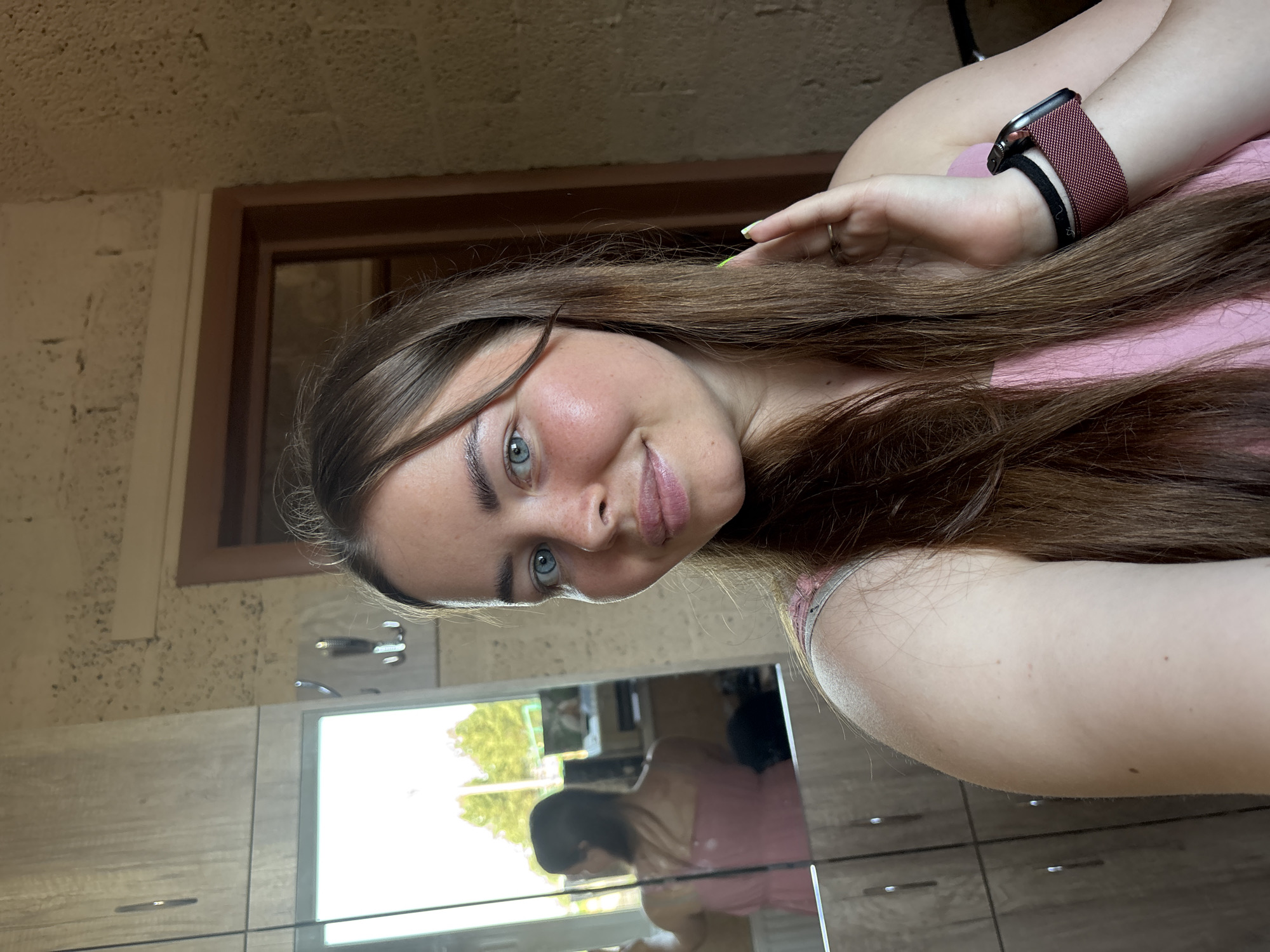 A young woman with long brown hair and blue eyes is posing for a selfie. She's wearing a pink top and an Apple Watch with a maroon band on her left wrist. Her left hand is raised near her hair. The background includes a window with a brown frame and a wall, potentially stone or with a textured finish. Below the window is a cabinet or storage unit with a reflective surface, showing a partial reflection of the woman. It is not possible to determine the location as Русский Вожой, Russia based on this image.