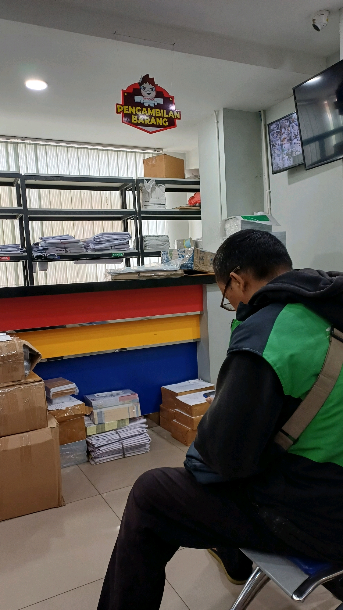 The image depicts the interior of what appears to be a courier or delivery service office in Cibinong, Indonesia. A man, seen from behind and side, is seated on a metal chair, wearing a black hoodie over a green vest and dark pants. He is looking down at a mobile device he is holding in his hands.
The environment is functional and organized, with multiple shelves laden with packages, boxes, and stacks of documents. The shelves are dark metal and are filled with items in various states of organization. Some items are neatly stacked, while others appear more haphazard. On the wall above the counter, a red and yellow hexagonal sign hangs, displaying text that appears to be Indonesian for "Package Pickup." To the right of this sign, a television screen is mounted on the wall, displaying a grid of surveillance camera feeds. The counter itself has distinct colored bands of red, yellow, and blue. In the foreground, several cardboard boxes are stacked on the floor. The floor is tiled in a light beige color. The lighting in the room appears to be from overhead fluorescent lights. The overall impression is that of a busy operational space within a logistics or delivery company.