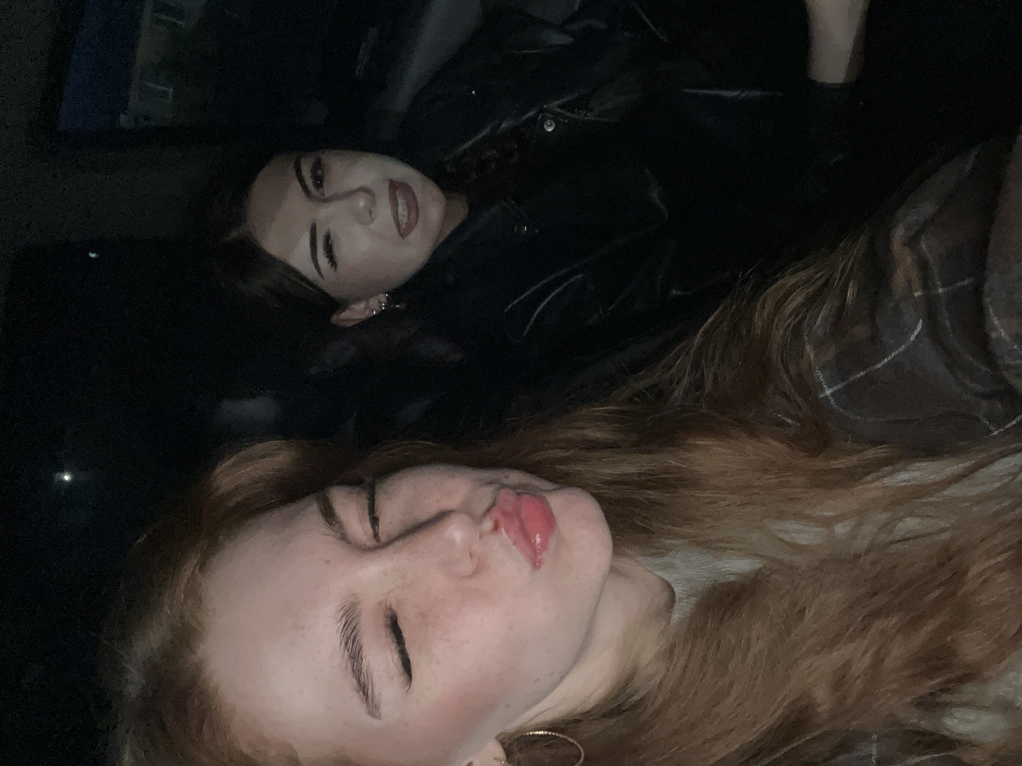 Two young women are captured in a selfie, taken in the interior of a vehicle at night, possibly in the city of Prudy, Russia. The woman in the foreground on the left has long, reddish-brown hair and is making a kissing face with her eyes closed. She is wearing a light-colored sweater and a hoop earring. To her right, the second woman is also making a face, sticking her tongue out slightly and winking. She has dark hair pulled back and is wearing a black leather jacket and small hoop earrings. The background is dark, with faint lights visible outside the vehicle, suggesting an urban or suburban environment. The overall lighting is dim, characteristic of an evening or nighttime photograph taken inside a car.
