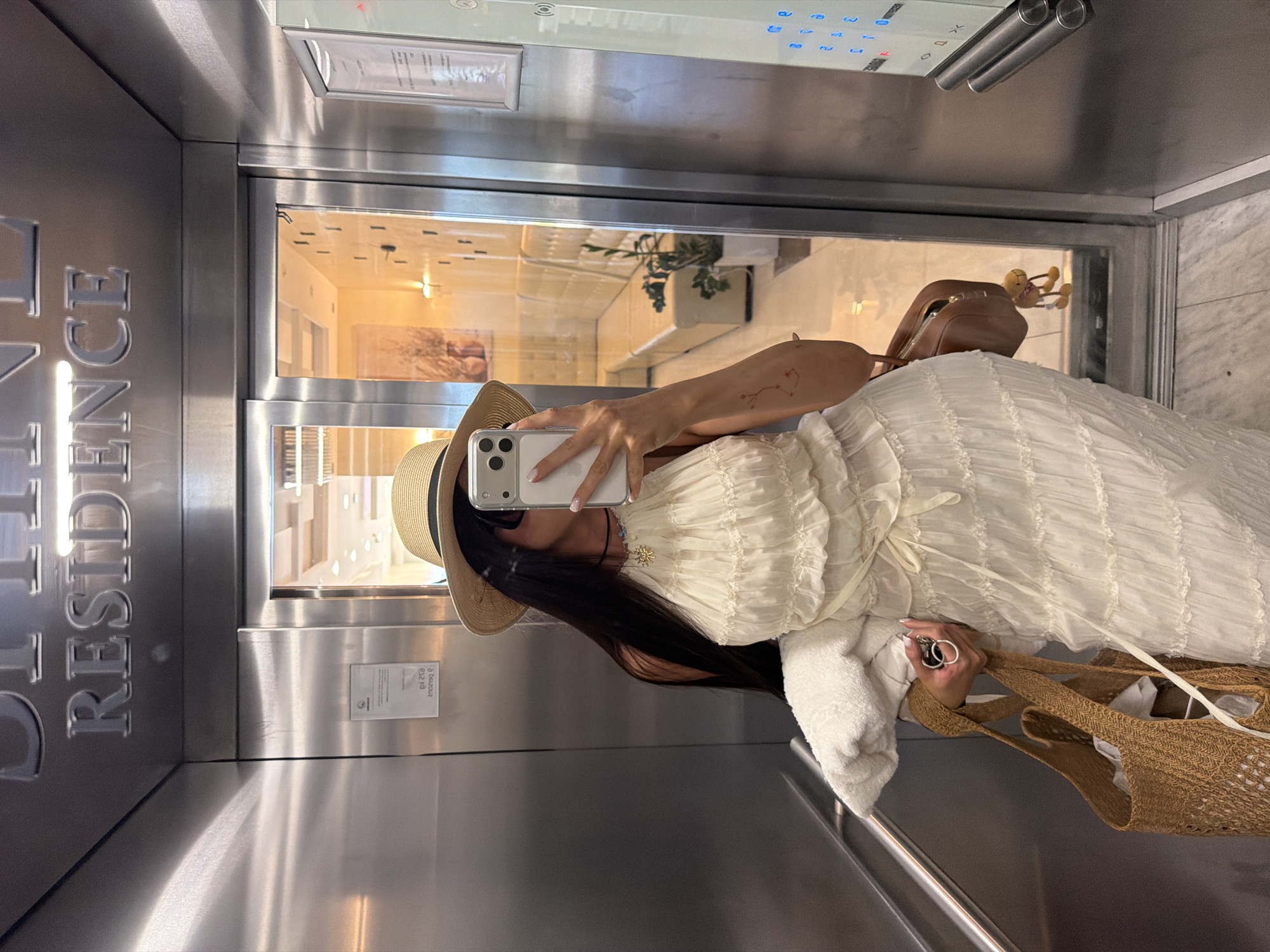 A woman with long, dark hair, wearing a straw hat, sunglasses, and a white tiered dress, stands in an elevator. She is holding a smartphone in her right hand, taking a selfie, and in her left hand, she holds a straw tote bag and keys. A white towel or wrap is draped over her left arm. The elevator walls are stainless steel, and above the elevator doors, silver lettering reads "RESIDENCE". The elevator doors are open, revealing a hallway with a seating area, plants, and decorative elements. The floor appears to be tiled. This scene is located in an elevator, likely in an accommodation building in the city of Boreti, Montenegro.
