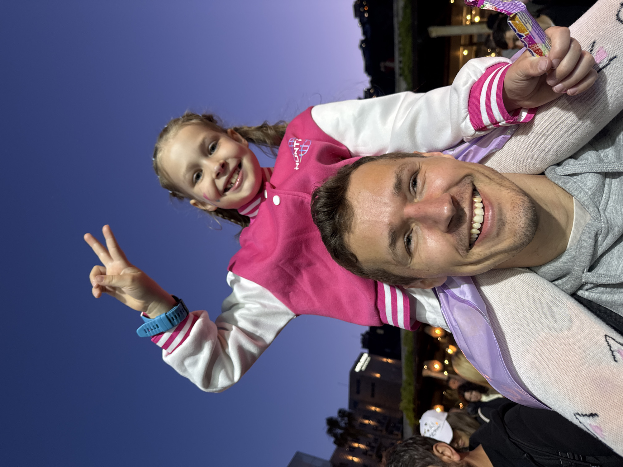 A man is carrying a young girl on his shoulders in an outdoor setting, likely in Budva, Montenegro. The girl is smiling and making a "peace" sign with her fingers, wearing a pink and white jacket, and white tights with an image on them. She also holds a wrapped candy bar in one hand. The man, who is also smiling, is wearing a gray sweatshirt.
The background includes other people, lights, and what appear to be buildings, indicative of a populated outdoor area. The sky is a deep blue, suggesting it is evening. The location is likely in Budva, Montenegro, given the style of architecture and the general atmosphere.