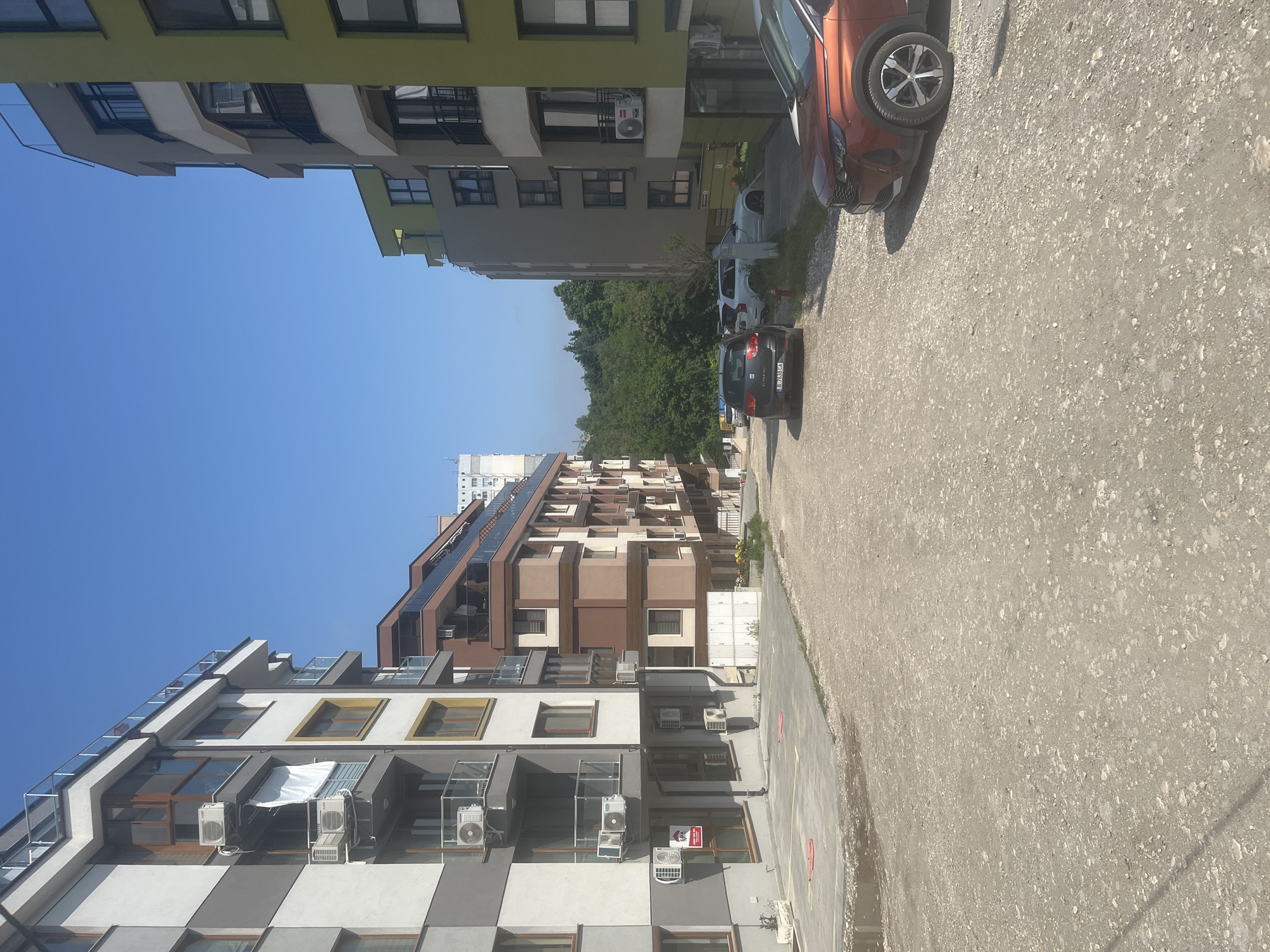 The image portrays a sunlit residential street scene, featuring multiple modern apartment buildings and parked vehicles under a clear blue sky. On the left, a multi-story building presents a distinctive grey and white checkered pattern with yellow window accents, adorned with several air conditioning units and balconies. In the mid-ground, another large building features a facade with various shades of brown, grey, and white, also equipped with balconies and air conditioning units. A third building with a green upper section and grey lower section is partially visible on the right. Lush green trees are visible in the mid-ground, situated between the buildings.
The immediate foreground consists of a rough, unpaved road or parking area covered in small stones. Further along, a section of paved road with faint yellow lines is visible. Several cars are parked, including the front section of an orange SUV in the foreground on the right, and a dark grey SEAT hatchback with the Bulgarian license plate "BZ 7539 KA" further down the road. Other white cars are parked in the background.
No explicit human activity or event is taking place; the scene captures a quiet, everyday urban environment. The bright, clear blue sky and strong illumination suggest it is mid-day with sunny weather. Visible text includes "ПРОДАВА" (FOR SALE) and "НАЕМ" (FOR RENT) on a red real estate sign attached to one of the buildings, along with partially visible phone numbers ("089..." and "0888 888...") and potentially a company name ("imoti"). The license plate "BZ 7539 KA" on the dark car indicates a Bulgarian registration, consistent with the Varna, Bulgaria, location context.