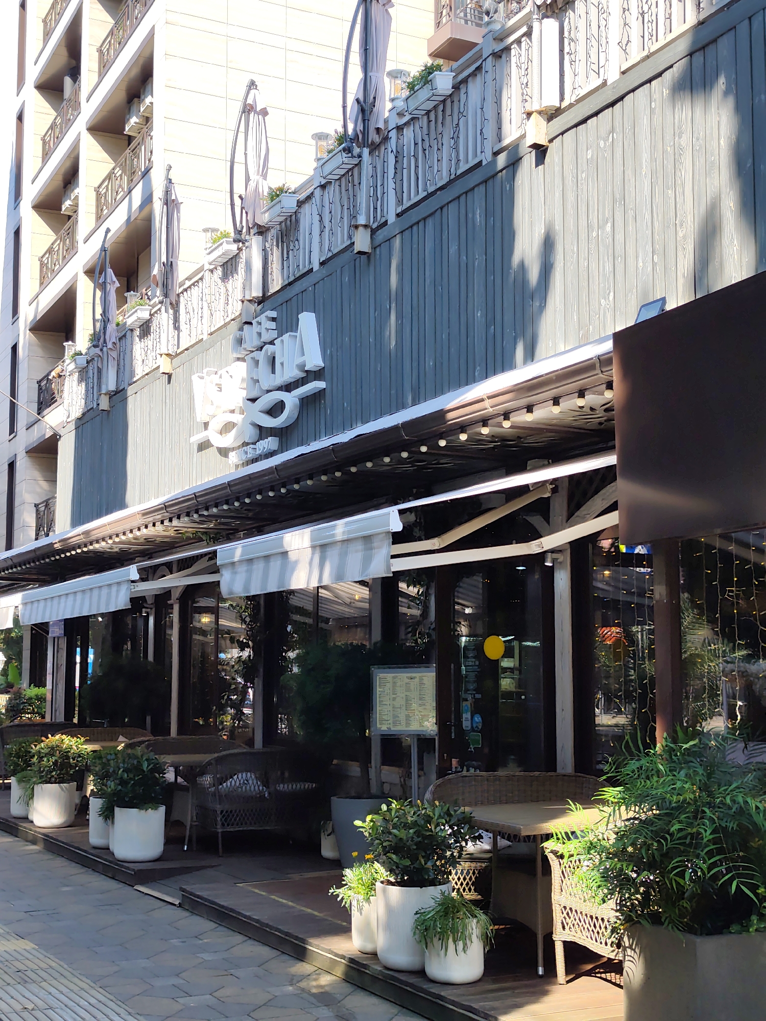 This image shows the exterior of a cafe named "The Chi" located on the sunny promenade of the city of Sochi, Russia. The cafe has a wooden facade with a sign featuring the cafe's name in white, stylized lettering. Outside the cafe, there are several tables and chairs set up on a wooden deck, with various potted plants providing greenery. The tables are made of wicker and wood, and some are covered with awnings. In the background, a modern building with balconies rises up. The promenade pavement is visible in the foreground. There are no people clearly visible in the scene.