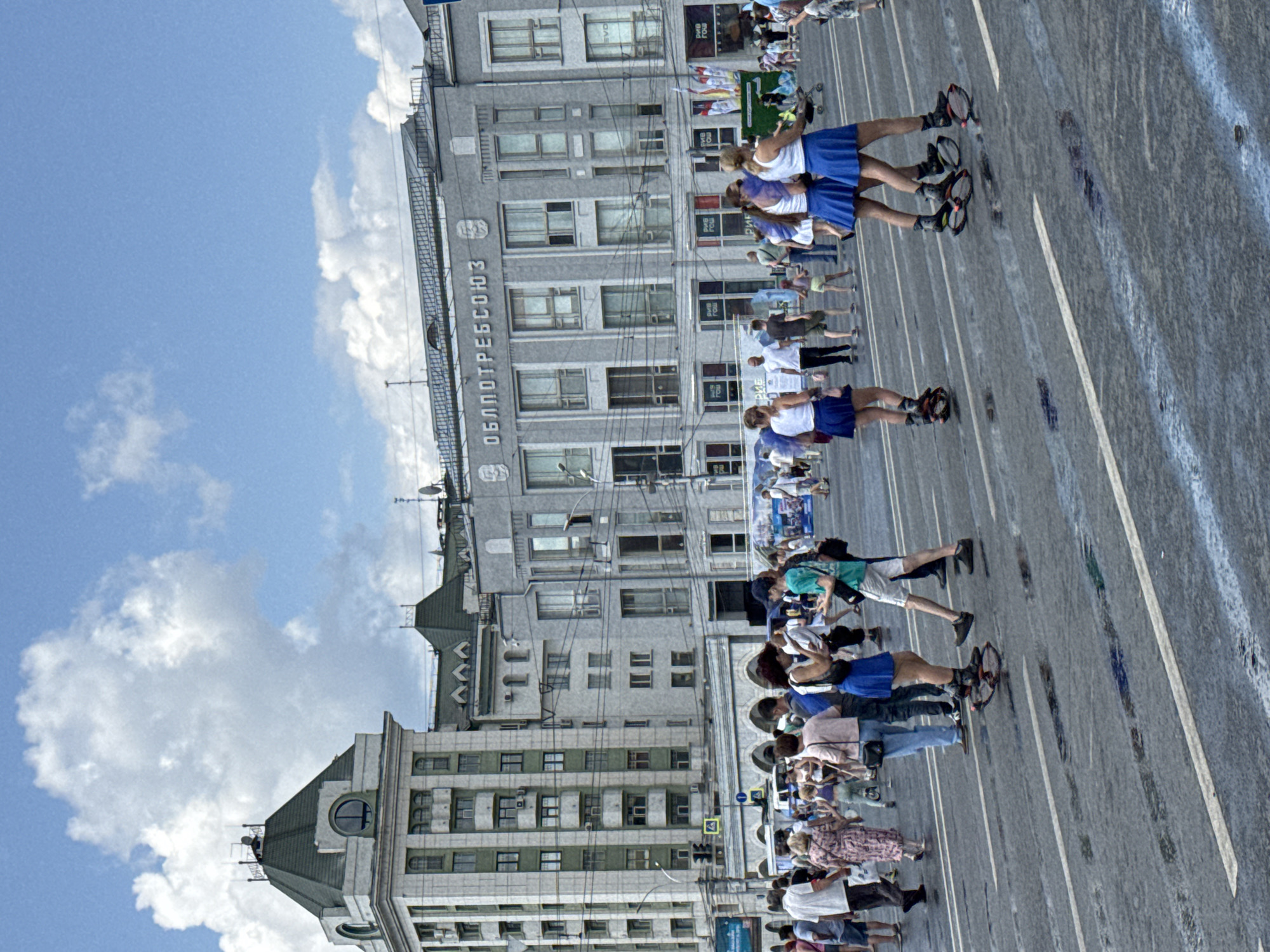 The image depicts a street scene in Novosibirsk, Russia, likely on a summer day with a partly cloudy sky. The foreground shows a wet asphalt road with white lane markings and reflective puddles. Several individuals are visible, some wearing distinct roller-skate-like devices on their feet, including three women in blue skirts and white tops in the mid-ground, and others scattered throughout the scene.
In the mid-ground, a crowd of people are walking on the street, some appearing to be spectators or participants in an event. Behind the crowd, several buildings form the backdrop. The most prominent building on the left is a multi-story structure with a green, mansard-style roof and a prominent clock tower. To its right, a long, grey building with numerous windows and the Cyrillic inscription "ОБЛПОТРЕБСОЮЗ" (Oblpotrebsoyuz) is visible. Further to the right, another grey building with a similar architectural style is present. Several banners and flags are also visible on poles in front of the buildings.
The overall impression is a public gathering or event taking place on a city street, with a mix of pedestrian activity and architectural elements characteristic of Russian urban landscapes. The presence of the roller-skate-like devices suggests a recreational or performance activity.