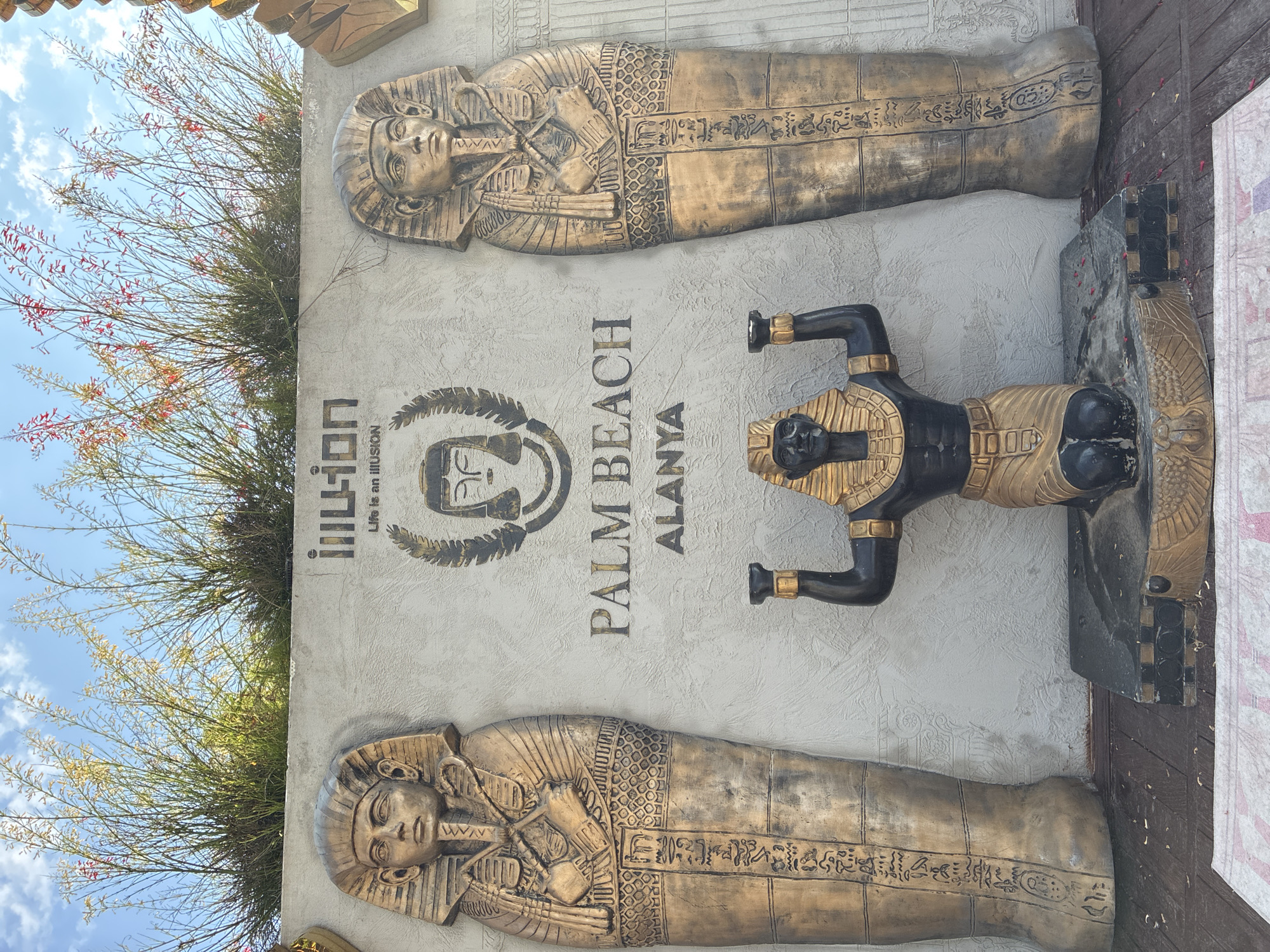The image shows an outdoor display with Egyptian-themed statues and lettering on a textured wall. Two large, golden sarcophagi figures stand on either side of the wall, each adorned with hieroglyphics. In the center, a black and gold statue of a pharaoh kneels, holding two staffs. Above the pharaoh statue, the word "ILLUSION" is written in gold, with the tagline "Life is an illusion" below it. Further down the wall, "PALM BEACH ALANYA" is inscribed in gold lettering. A lush green plant with small red flowers grows above the wall, with a blue sky and scattered clouds in the background. The display is situated on a wooden deck, with a patterned rug partially visible in the foreground. This is likely part of the "Illusion" themed attraction at Palm Beach in Alanya, Turkey, a coastal resort city known for its historical sites and beaches.