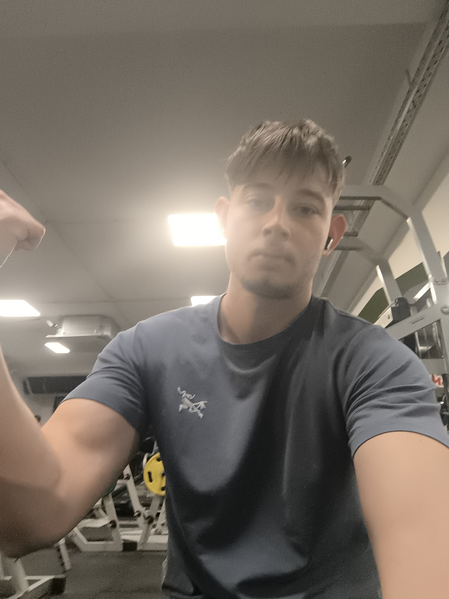 A young man is taking a selfie in a gym. He is wearing a gray T-shirt with a logo on the left side of his chest and an earbud in his right ear. He is flexing his left bicep. In the background are various pieces of gym equipment, including weight machines and racks. The gym has fluorescent lighting. The location is a gym, but the city of Sofia, Bulgaria cannot be determined.
