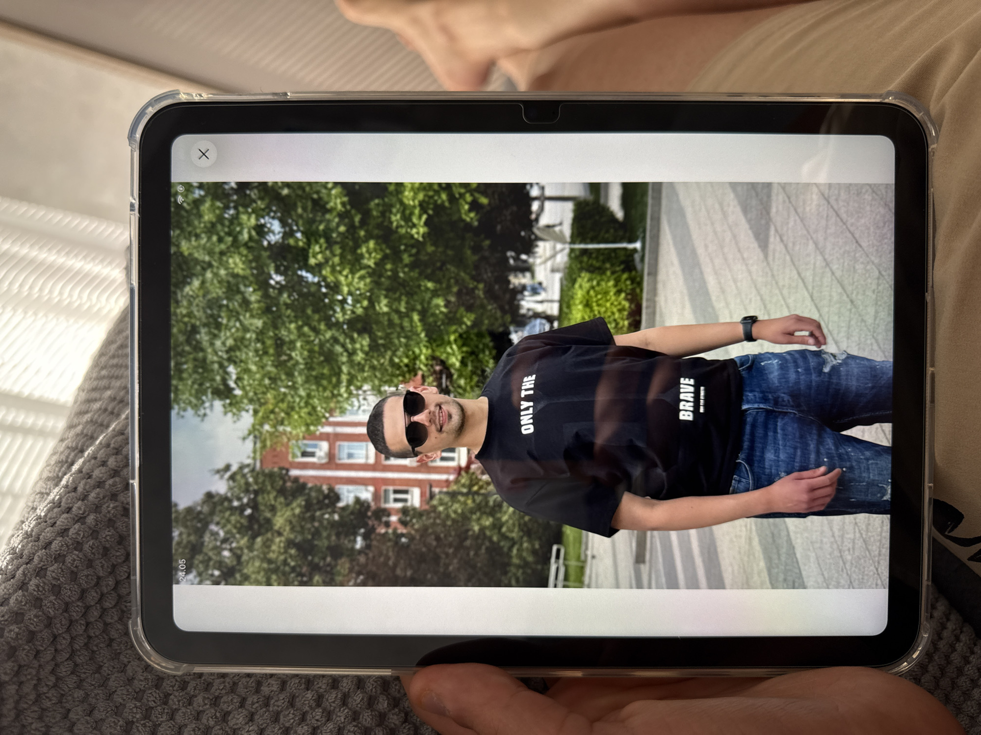 A photograph shows an iPad displaying an image of a man. The man is wearing a black t-shirt with the words "ONLY THE BRAVE" printed on it, blue jeans, and sunglasses. He appears to be standing on a sidewalk in front of trees and a red brick building. The date "24.05" appears on the left side of the iPad screen. The man in the picture is standing in the city of Lovech, Bulgaria.