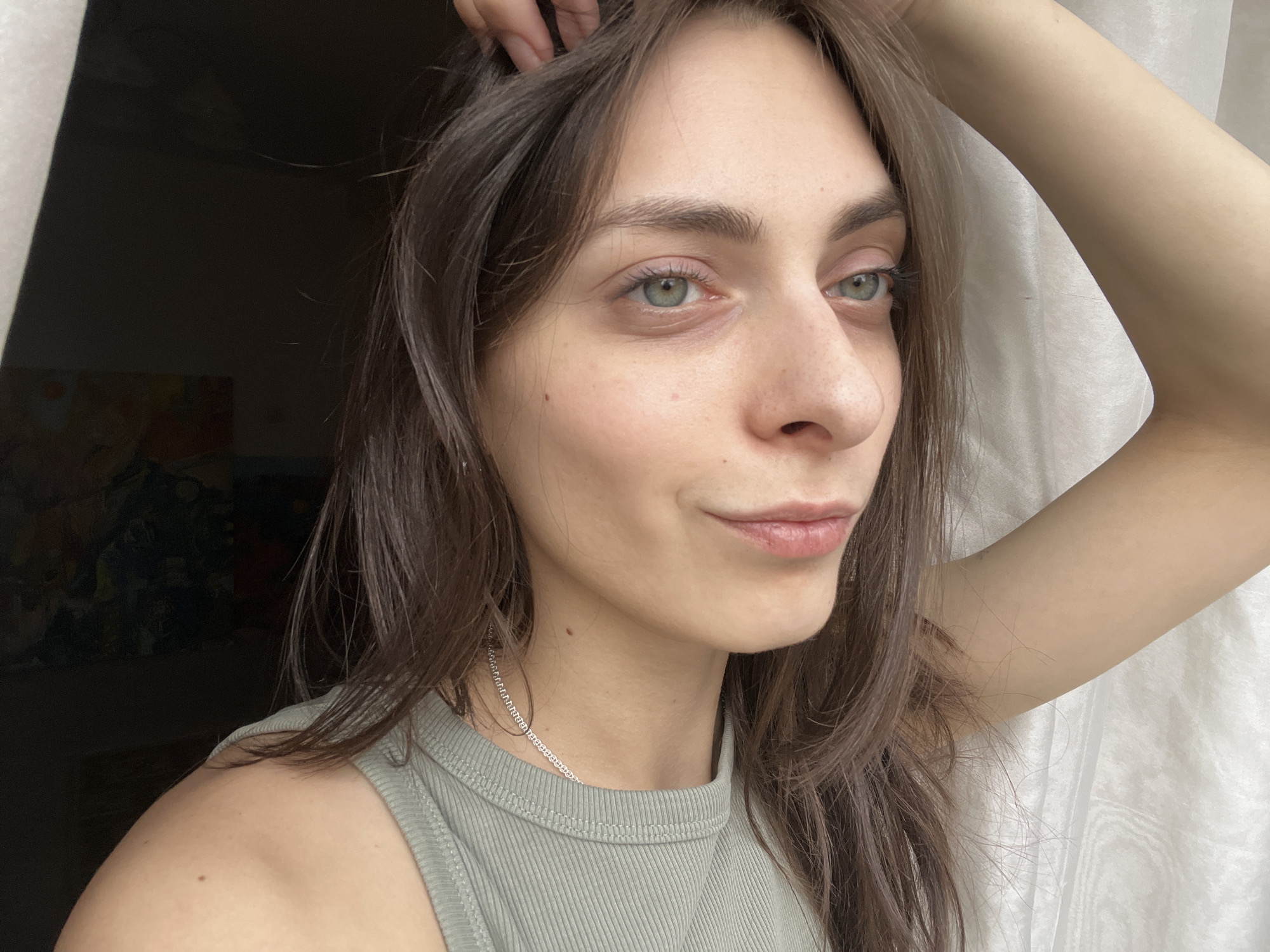 A close-up portrait captures a young woman indoors in Krasnoyarsk, Russia. She has long, dark brown hair, which appears casually styled, with her right hand gently running through the strands near her scalp. Her fair complexion shows natural freckles across her nose and upper cheeks, and a small, round mole is visible on her neck and another on her left shoulder. Her light green or hazel eyes are directed slightly to her right, away from the camera, and her lips form a subtle, soft smile or smirk, conveying a calm or thoughtful expression.
She is dressed in a light olive-green ribbed tank top, and a thin silver chain necklace adorns her neck. The lighting is soft and natural, suggesting daytime, likely coming from a window. The background on the left is dark, hinting at a wall or a darker interior space, with a dark, abstract painting or print visible in the lower left corner. On the right side of the frame, sheer white or off-white curtains are visible, confirming the presence of a window nearby. Her pose is relaxed, with her right arm raised, and her body angled slightly. There is no visible text in the image.