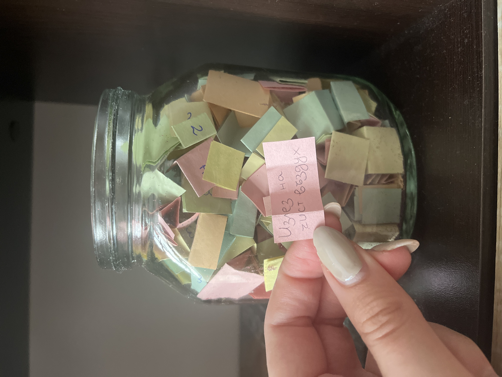 A hand holds a small, pink piece of paper with writing in Cyrillic lettering near a glass jar filled with similarly colored small, square pieces of paper, some also with Cyrillic lettering. The jar sits on a dark brown surface. The context of the location is not discernible from the image to determine if it is in the city of Varna, Bulgaria.