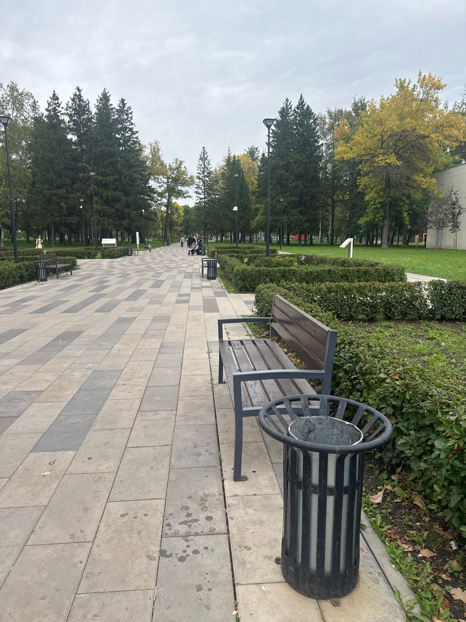 The image depicts a public park walkway in Ufa, Russia, characterized by a cloudy autumn sky and lush greenery. The path is paved with light-colored square and rectangular tiles arranged in a patterned fashion, bordered by manicured hedges on the right. Several benches with dark wooden seats and metal frames are positioned along the walkway, accompanied by cylindrical trash receptacles. The trees in the background display a mix of green and yellow foliage, indicative of the autumn season. A few individuals are visible in the distance, some pushing strollers, suggesting a leisurely atmosphere. The overall scene portrays a well-maintained urban park environment.