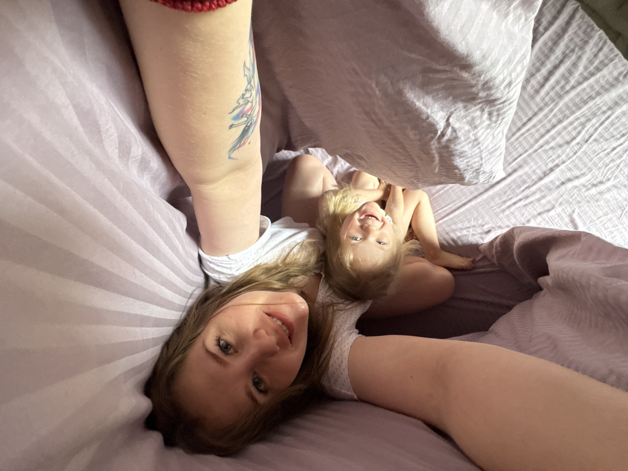 A light-skinned woman with long blonde hair lies on a bed with lavender-colored striped sheets. She is smiling at the camera. A young blonde-haired child with light skin is under the covers, also smiling at the camera. A woman's arm with a tattoo is visible near the top left of the frame. The location appears to be indoors. The city of Gornyi (Dvurechenskoe S/P), Russia, is not discernible from the image.