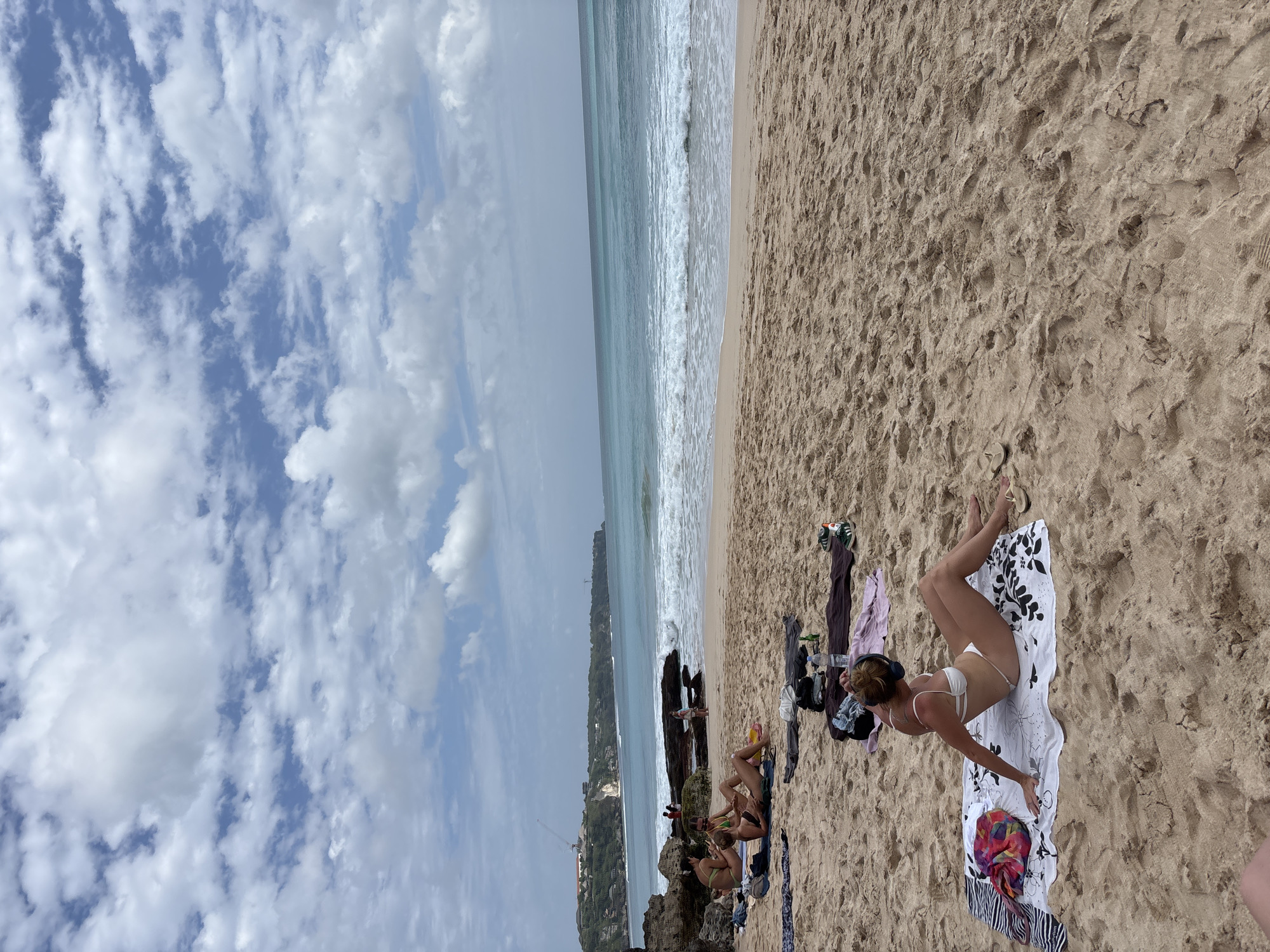 This image depicts a scene at a beach in Kuta Selatan, Indonesia, likely Padang Padang Beach or a similar location known for its scenic beauty and surf. The sand is a light brown, with footprints and patterns indicative of a busy beach.
In the foreground, a woman in a white bikini is seated on a towel, facing away from the camera and towards the ocean. She has headphones on and her left arm is raised slightly. A colorful sarong or scarf lies to her left. Further back, closer to the water's edge, several other people are sunbathing and relaxing on towels. One group of three women are sitting and conversing, one wearing a green bikini, another a brown one, and the third in a black bikini. Another individual is lying down on a towel.
The ocean is a vibrant turquoise, with moderate waves breaking on the shore, creating white foam. In the distance, a cliff face runs along the coastline, with buildings and a construction crane visible on top of the cliffs, suggesting development in the area. The sky is partly cloudy, with white cumulus clouds scattered across a blue expanse. The overall atmosphere is one of a leisurely day at the beach, with people enjoying the sun and the sea.