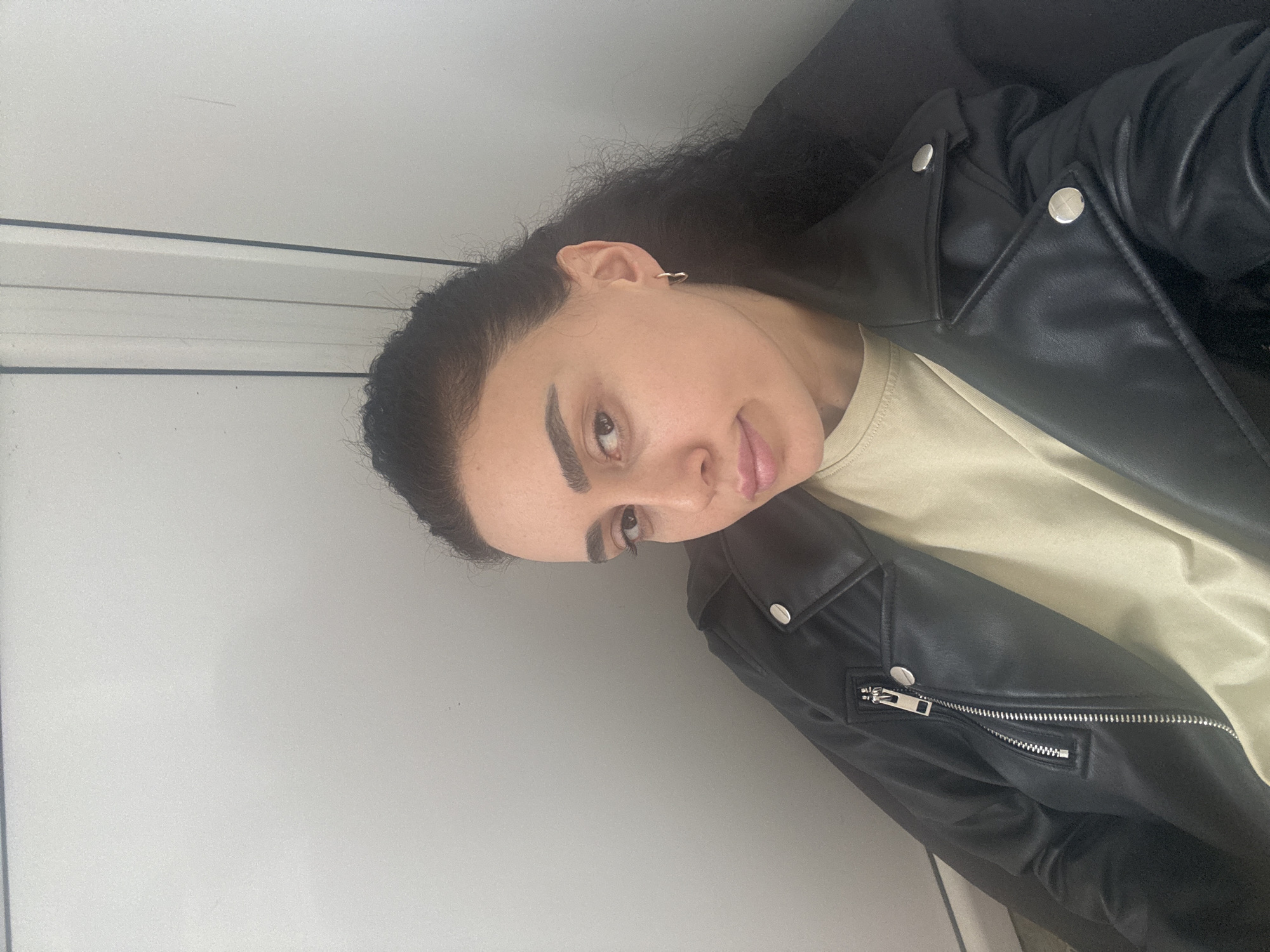 A woman is positioned against a white wall. She is wearing a light-colored shirt and a black leather jacket. Her dark curly hair is pulled back. She has earrings in her ear. No location in Tbilisi, Georgia is discernible from the image.