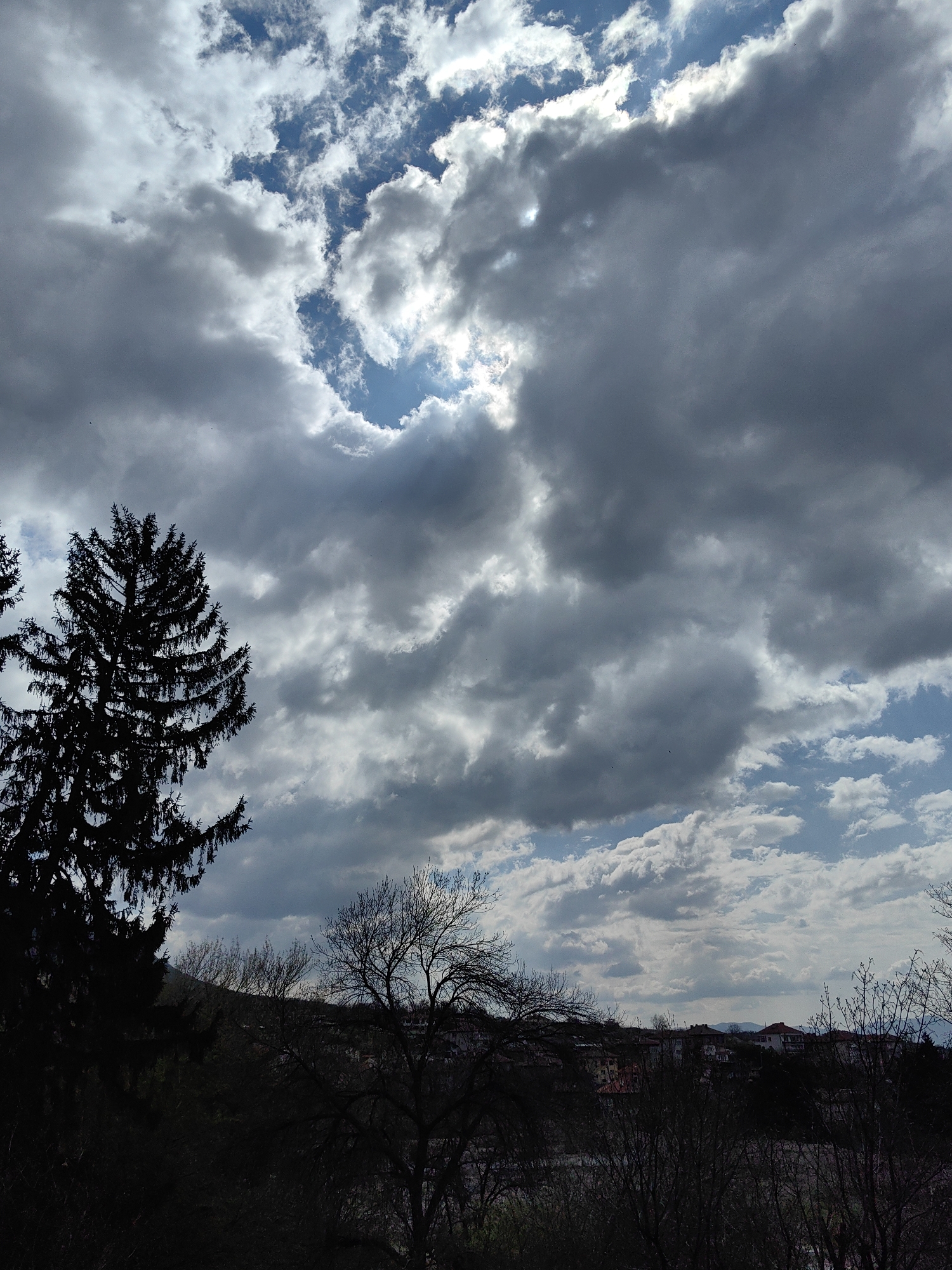 The image captures a partially cloudy sky over Karlovo, Bulgaria. Dominating the view is a sky filled with a mix of dark, heavy clouds and patches of blue sky with smaller, white clouds. The sunlight peeks through a hole in the clouds. In the foreground, the silhouettes of several trees stand against the skyline. The town itself is visible in the middle distance, showing buildings with red roofs and light-colored walls. In the far distance, one can make out the faint outline of mountains.