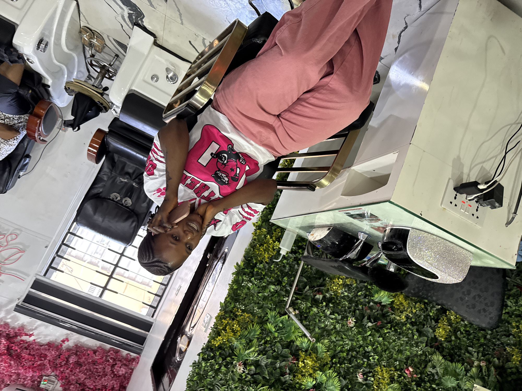 A young woman sits in a salon chair, possibly in Lugbe, Nigeria. She wears a white t-shirt with a pink logo and pink sweatpants. The salon features manicure stations, one with a white and silver embellished object, and a wall with artificial foliage. There is a window with blinds, a pink floral arrangement, and pedicure stations nearby.