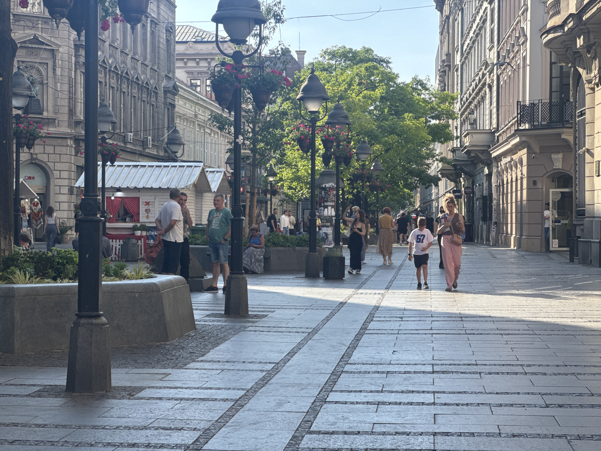 The image depicts a wide, pedestrianized street in Belgrade, Serbia, likely Knez Mihailova Street, given its architecture and lively atmosphere. The street is paved with large, grey stone slabs and narrow, darker stone borders. Sunlight casts distinct shadows across the pavement, indicating a bright, sunny day, likely in the late afternoon or early evening.
Along both sides of the street are ornate, multi-story buildings with classical architectural elements, including detailed facades, balconies with iron railings, and large windows. Numerous trees with lush green foliage are interspersed along the street, providing shade and softening the urban landscape. Decorative lampposts with hanging flower baskets are strategically placed, adding to the street's charm.
Several people are visible walking in both directions, suggesting a busy commercial and social hub. Some are casually strolling, while others appear to be engaged in shopping or conversation. A few individuals are standing or sitting near small kiosks or shops, further indicating commercial activity. Among the people, there's a clear mix of ages and attire, with some dressed in summer clothing, such as shorts and t-shirts, and others in more formal wear.
In the foreground, a large, grey stone planter with green plants is prominent, framing the left side of the image. A dark lamppost stands next to it. The overall impression is one of a vibrant, well-maintained urban environment where people are enjoying a pleasant day out.