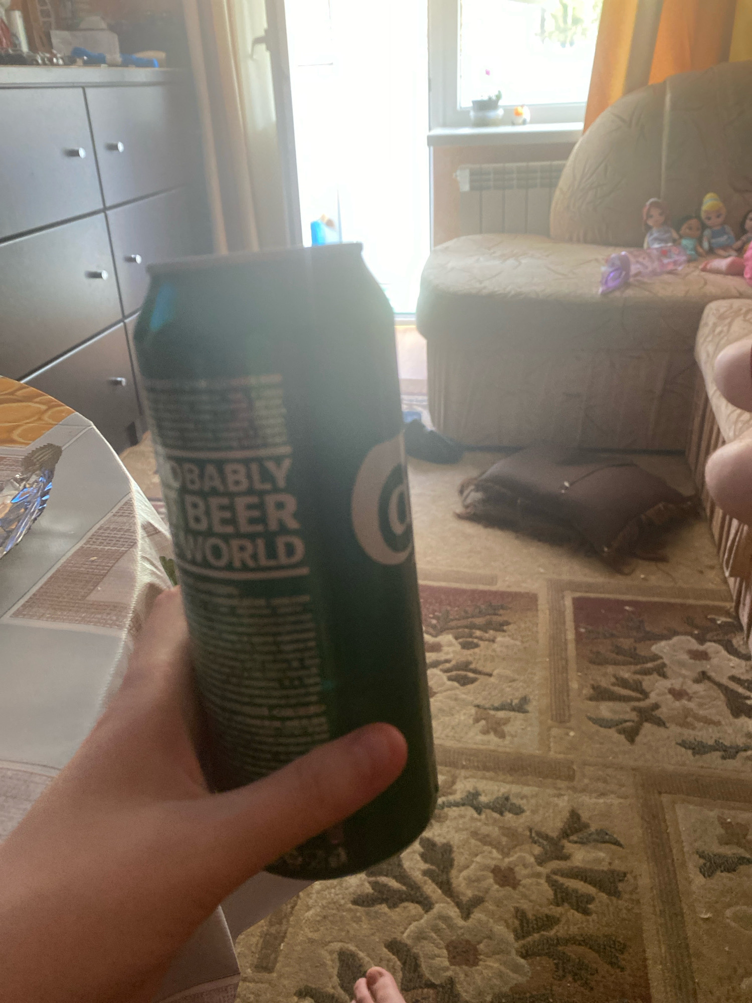 An indoor scene shows a hand holding a dark green beer can in the foreground. The can has text printed on it, partially visible as "ROBABLY BEER WORLD". The background reveals a room with a light-colored, plush sofa with several dolls seated on it. To the left of the sofa, a radiator is visible below a window. Further to the left, a dark brown dresser with several drawers is present. The floor is carpeted with a patterned rug in shades of brown and beige. The lighting in the scene suggests it is daytime, with natural light streaming in from the window. The overall setting appears to be a living room.