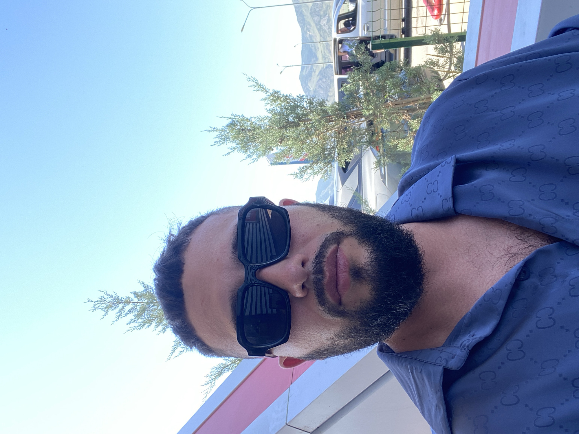 A man with dark hair and a beard wearing black sunglasses and a dark blue shirt with a repeating pattern is taking a selfie outdoors. He is positioned in the foreground, with a bright blue sky and distant mountains in the background. Several evergreen trees are visible, along with parts of vehicles and a light-colored structure with pink and white stripes. The scene appears to be in a somewhat arid, possibly mountainous region. The man is looking directly at the camera.