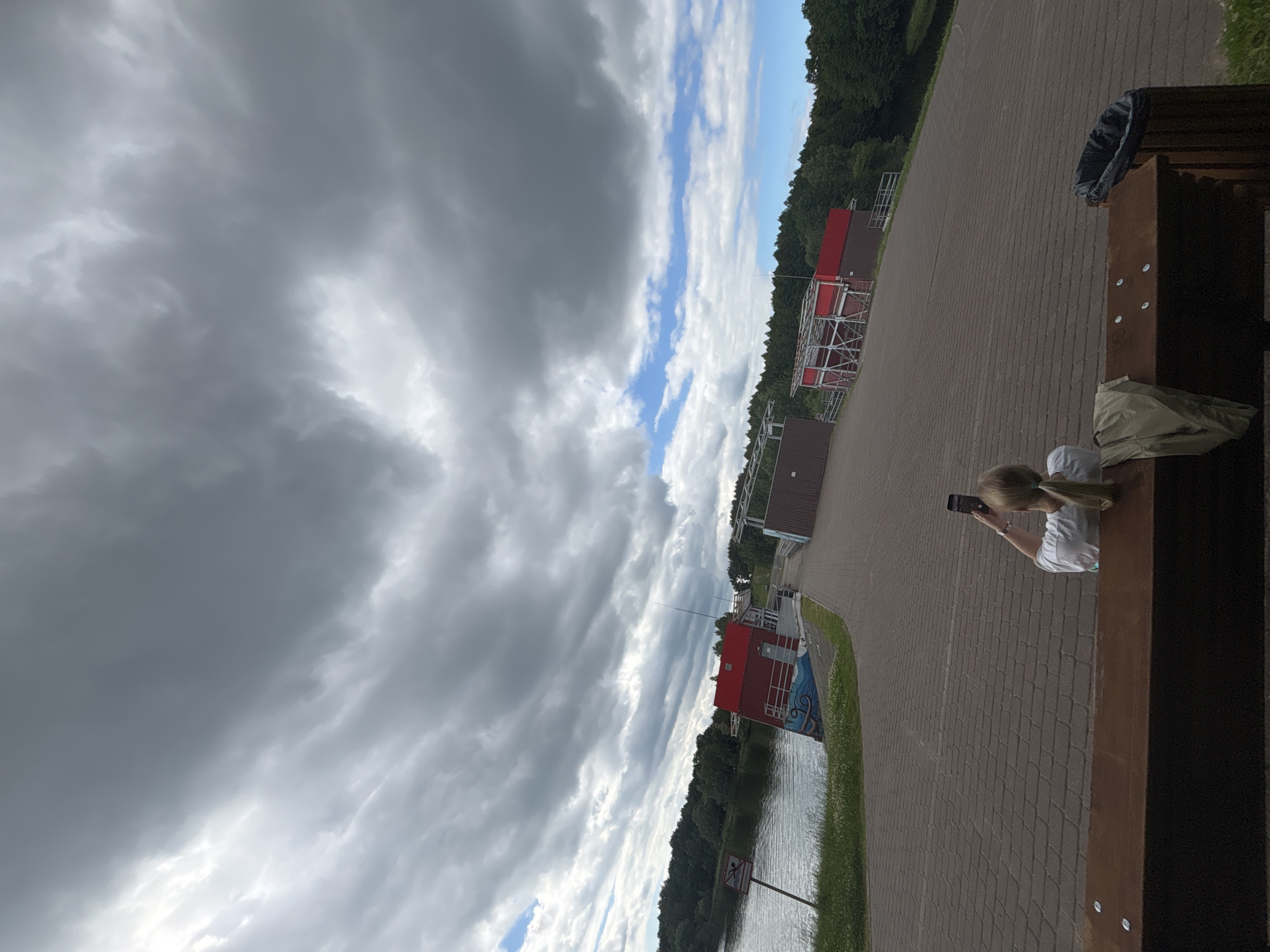 The image shows a scenic view of a lake and a building with red roofs, set against a backdrop of a cloudy sky. In the foreground, a woman is sitting on a bench and taking a picture with her phone. The location is identified as Minsk, Belarus.