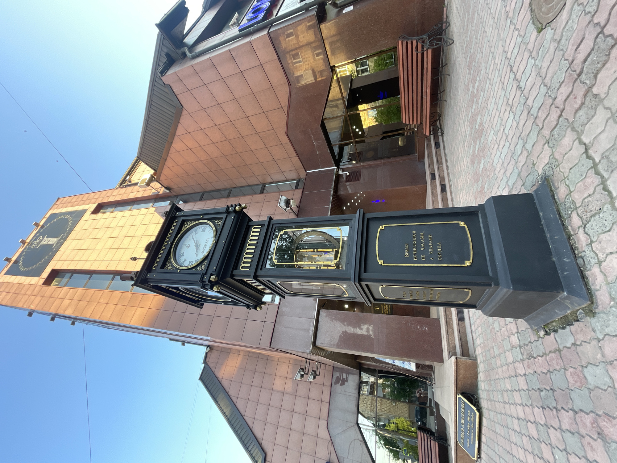 The image showcases an ornate, black and gold street clock standing in a pedestrian area, likely a square, in Krasnoyarsk, Russia. The multi-faced clock displays Roman numerals on one dial and reveals its intricate internal gears through a glass panel on another side. A prominent golden plaque on the clock's base features the Cyrillic quote: "ВРЕМЯ ИСЧИСЛЯЕТСЯ НЕ ЧАСАМИ, А УДАРАМИ СЕРДЦА," which translates to "Time is measured not by hours, but by heartbeats."
Behind the clock, a modern building with a reddish-brown paneled facade and reflective glass sections rises against a clear blue sky. The building's upper stories display signage with the words "IMPERIAL" and "ИМПЕРІАЛЬ." Reflections in the glass show parts of surrounding architecture and trees. A wooden bench with a dark metal frame is situated near the building's entrance. The ground is paved with patterned, light-colored interlocking bricks. A small ground-level plaque indicates the location as "ПЛОЩАДЬ ВЛЮБЛЕННЫХ" ("Lovers' Square") and references a date "дарыта 17 октября 200" (gifted October 17, 200...). The scene is captured on a bright, sunny day, with no people visible, focusing on the urban architectural elements and the decorative clock.