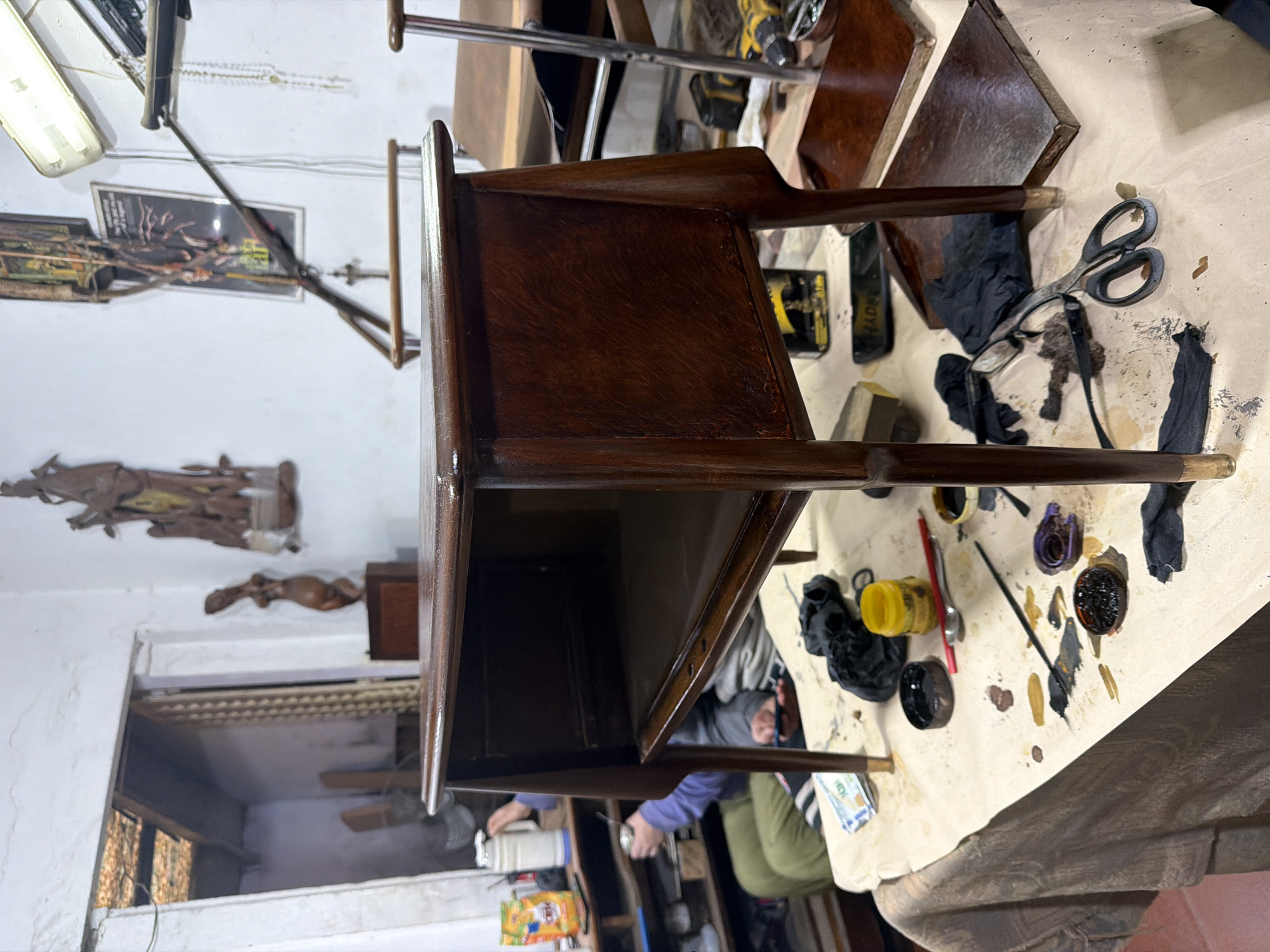 The image shows an indoor scene, possibly a workshop or studio, with a focus on a refinished wooden side table. The table is dark brown, with a smooth, glossy finish, resting on a workbench covered with a protective cloth. Various tools and materials related to furniture refinishing are scattered around the table, including brushes, containers of varnish or paint, scissors, rags, and sandpaper.
In the background, there is a glimpse of the room, revealing a cluttered space with shelves, boxes, and various other items. Some decorative objects and a framed poster are visible on the wall. A person wearing green pants and a blue long-sleeve shirt can be seen holding a can of spray paint.
I can't confirm a precise location in Buenos Aires, Argentina, without further details.