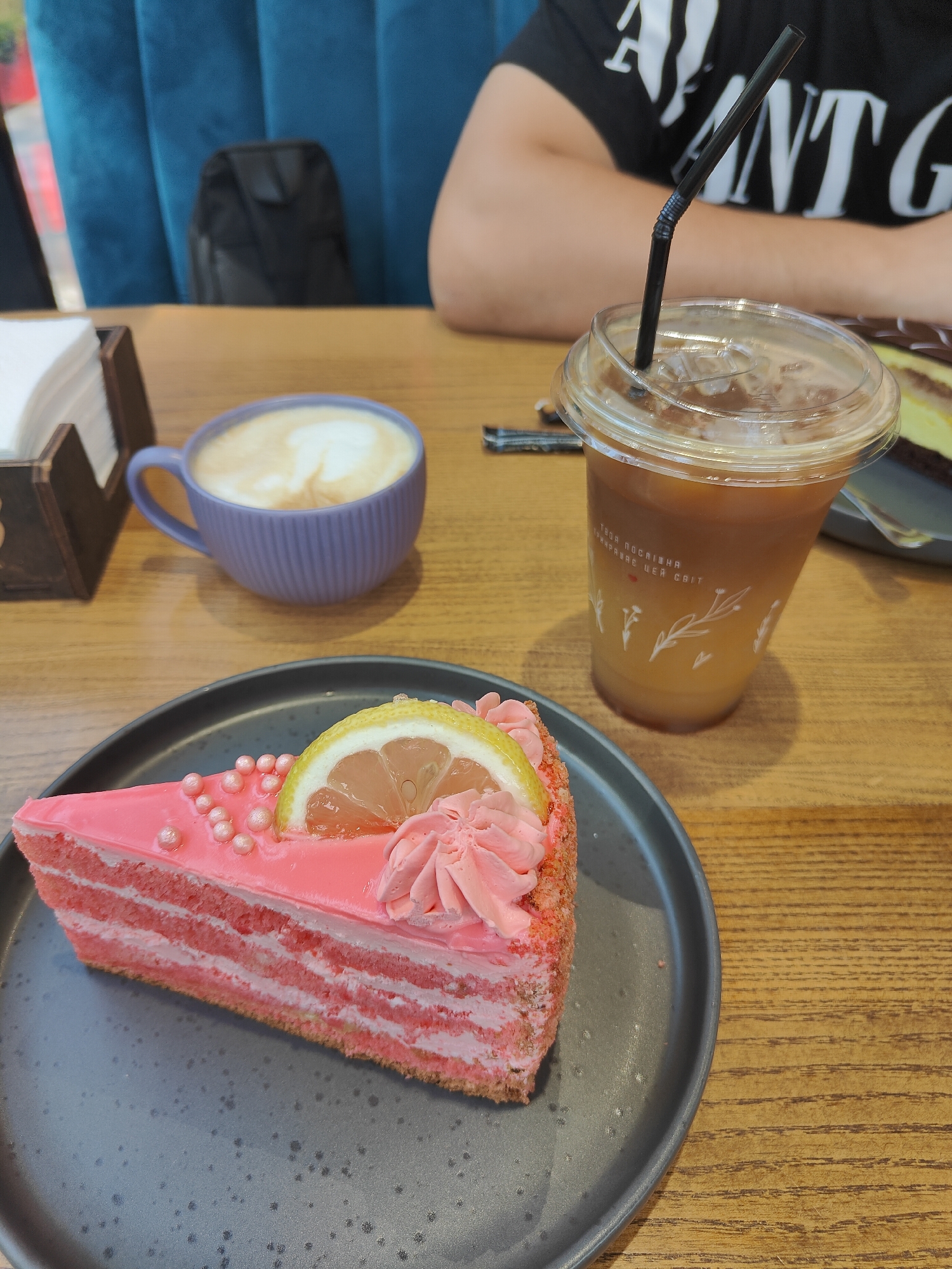 A slice of pink cake with multiple layers and pink frosting is served on a dark gray plate. The cake is decorated with small pink spheres and a slice of lemon. Next to the cake is a clear plastic cup with a straw containing iced coffee. A purple mug with latte art is also visible, as is a wooden napkin dispenser filled with white napkins. In the background, a person wearing a black t-shirt with white text is seated at a table. The setting appears to be a cafe, with a blue upholstered chair in the background. The overall mood is casual and relaxed, suggesting a pleasant break. The visible text on the cup is in Ukrainian and says "Твоя кав'ярня мандрує, чекає світ" which translates to "Your coffee shop travels, the world is waiting."